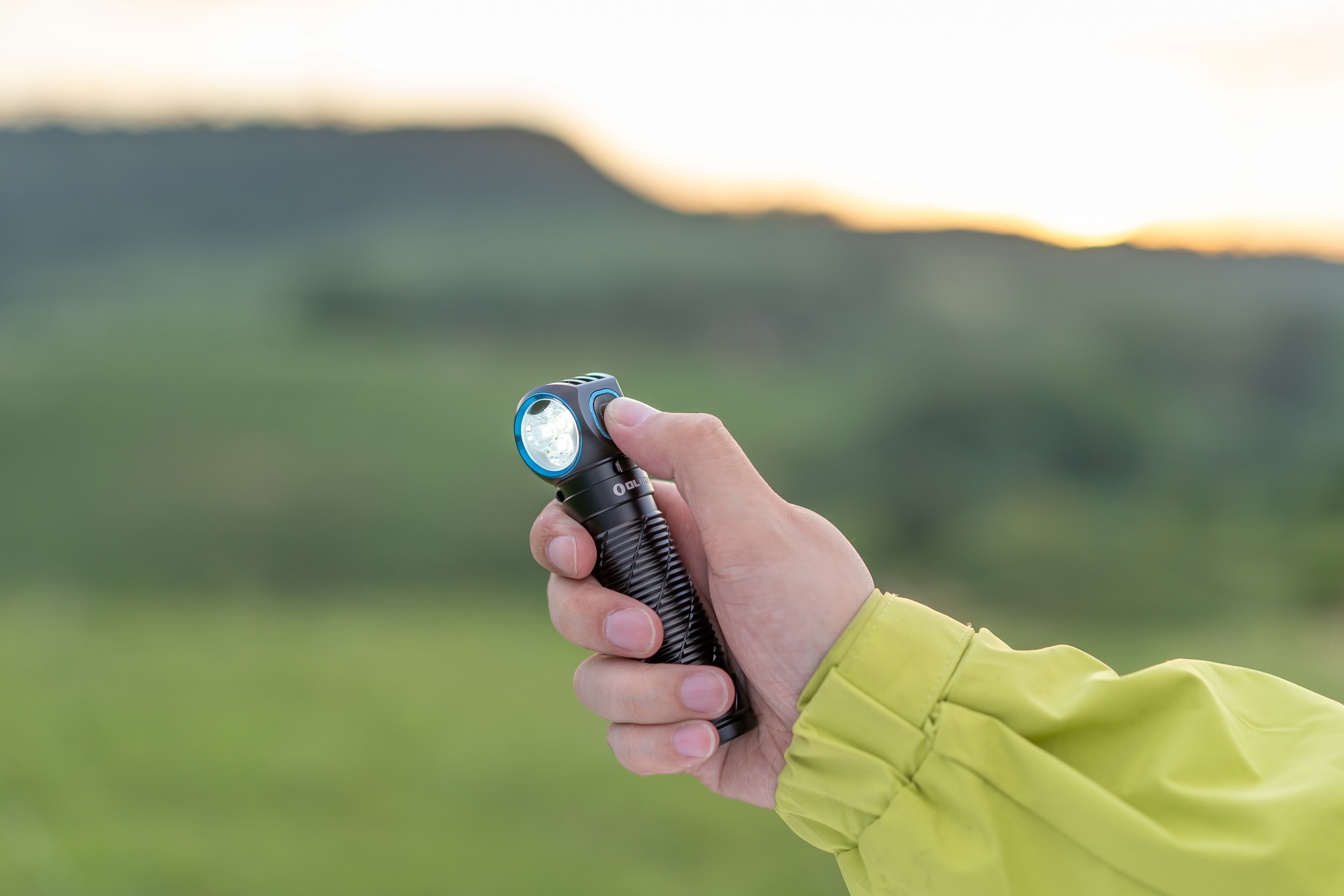 Hand holding an illuminated Olight headlamp outdoors at dusk, with a blurred natural landscape and sunset glow in the background.