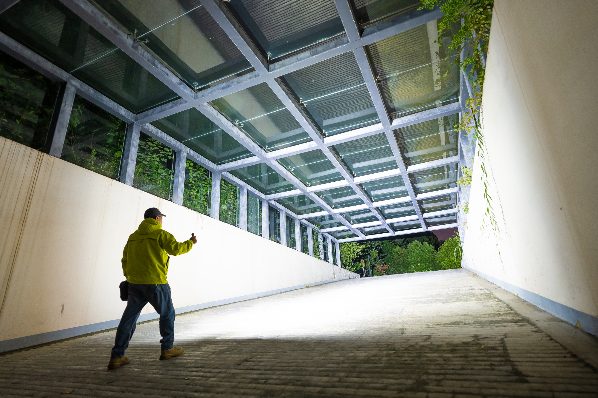 Man using a powerful high-lumen LED flashlight to illuminate a modern glass-roofed walkway at night, showcasing a bright light beam and outdoor security lighting.