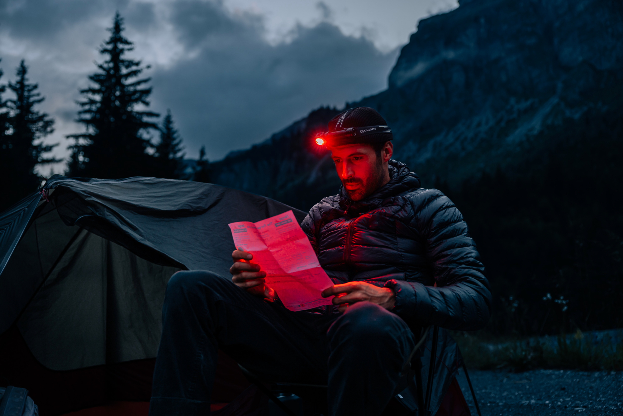 Man with red headlamp reading a map next to his tent in the mountains at night, planning a hike or outdoor adventure.