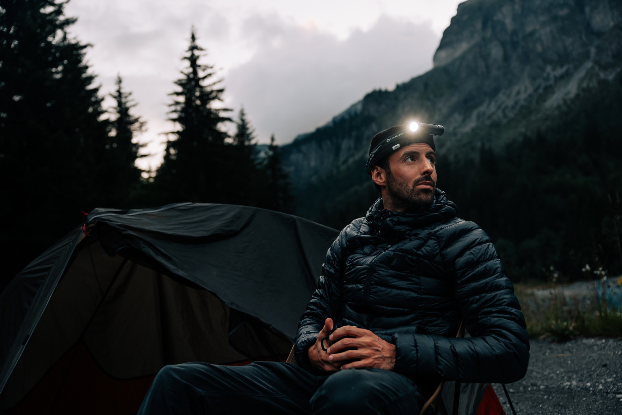 Man with Olight headlamp and dark jacket sitting by tent in mountain forest. Evening camping scene, outdoor adventure.