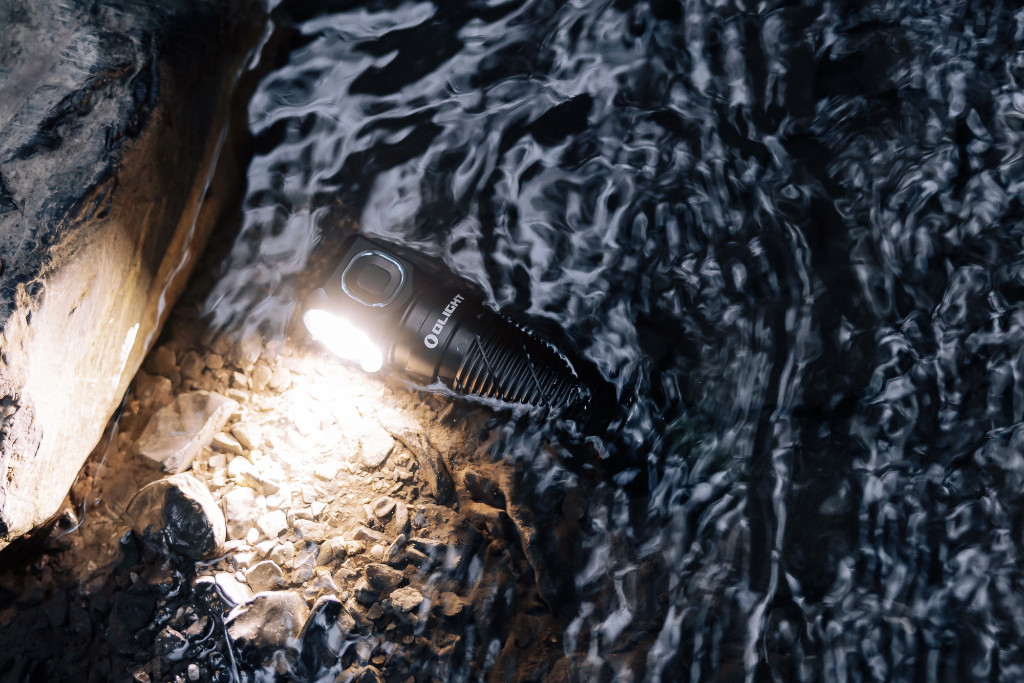 Olight waterproof tactical flashlight submerged in a stream, its bright beam illuminating wet rocks and rippling water.