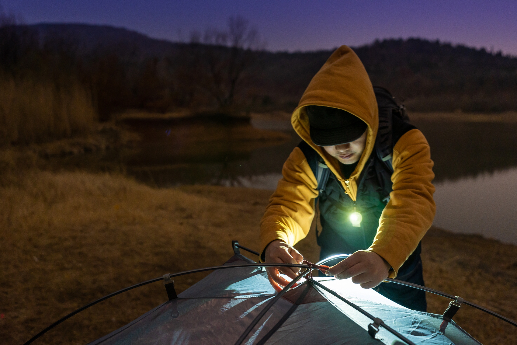 Un homme en sweat à capuche jaune monte sa tente de camping au bord d'un lac à la tombée de la nuit, éclairé by une lampe frontale.