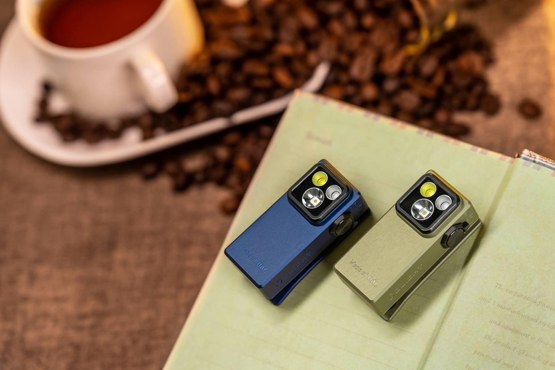 Dos linternas LED compactas, una azul y una verde oliva, sobre un libro abierto, con granos de café y una taza de café en el fondo.