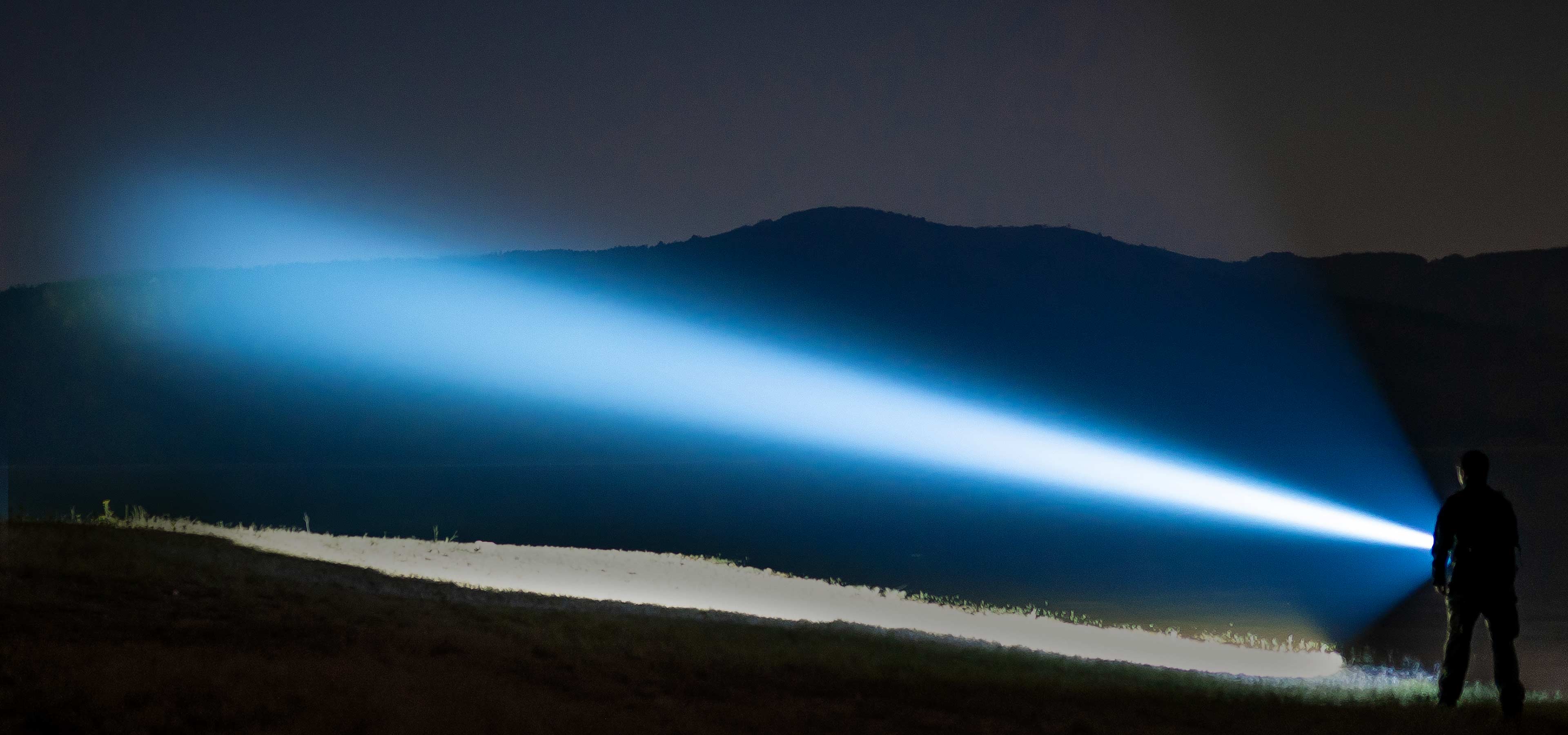 Persona con potente linterna proyectando un brillante haz de luz azul a través de un paisaje montañoso oscuro en la noche.
