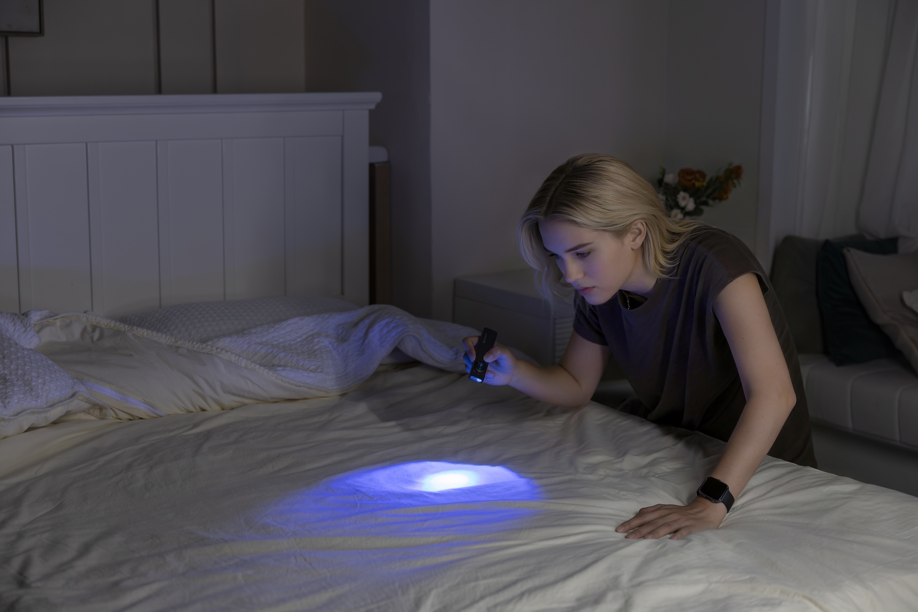 Young woman using a portable UV light flashlight to inspect white bed sheets for stains and cleanliness in a dark bedroom.