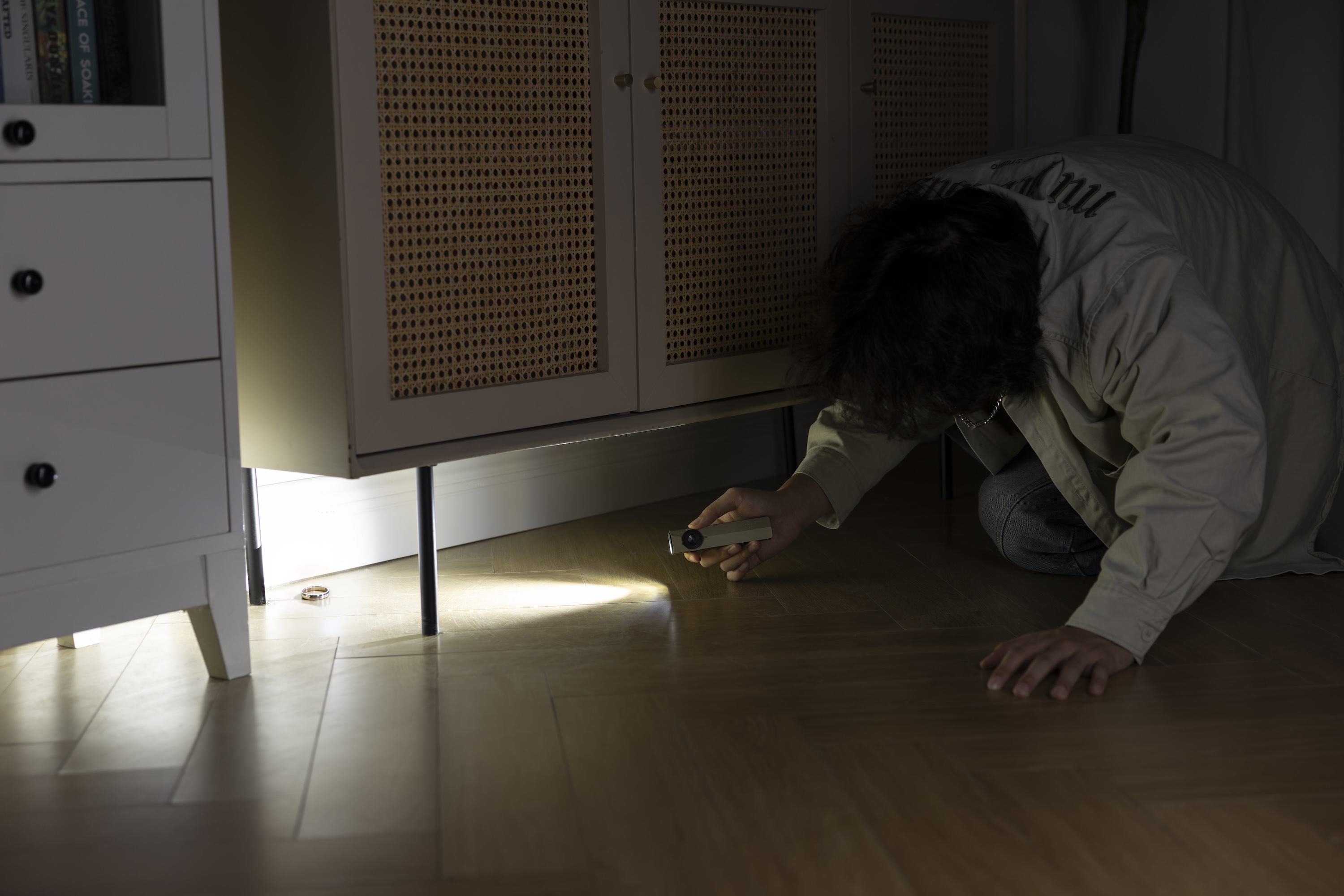 A person using a slim portable flashlight to search for a lost ring under a rattan cabinet on a wooden floor, demonstrating a compact light for finding items in dark corners.