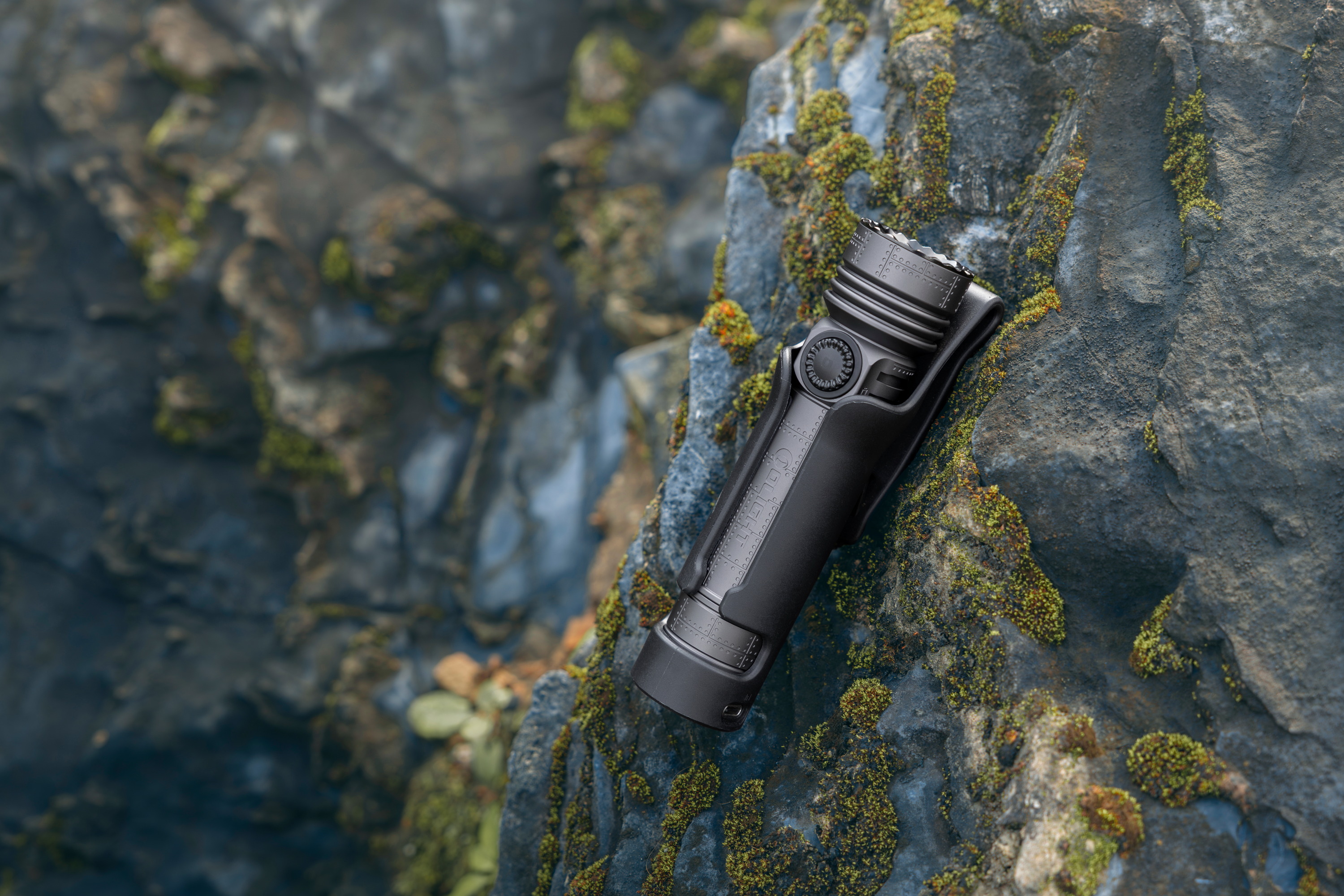 Linterna t&aacute;ctica militar negra en su funda, colocada sobre una roca natural con musgo h&uacute;medo en un entorno de monta&ntilde;a para actividades al aire libre y supervivencia.