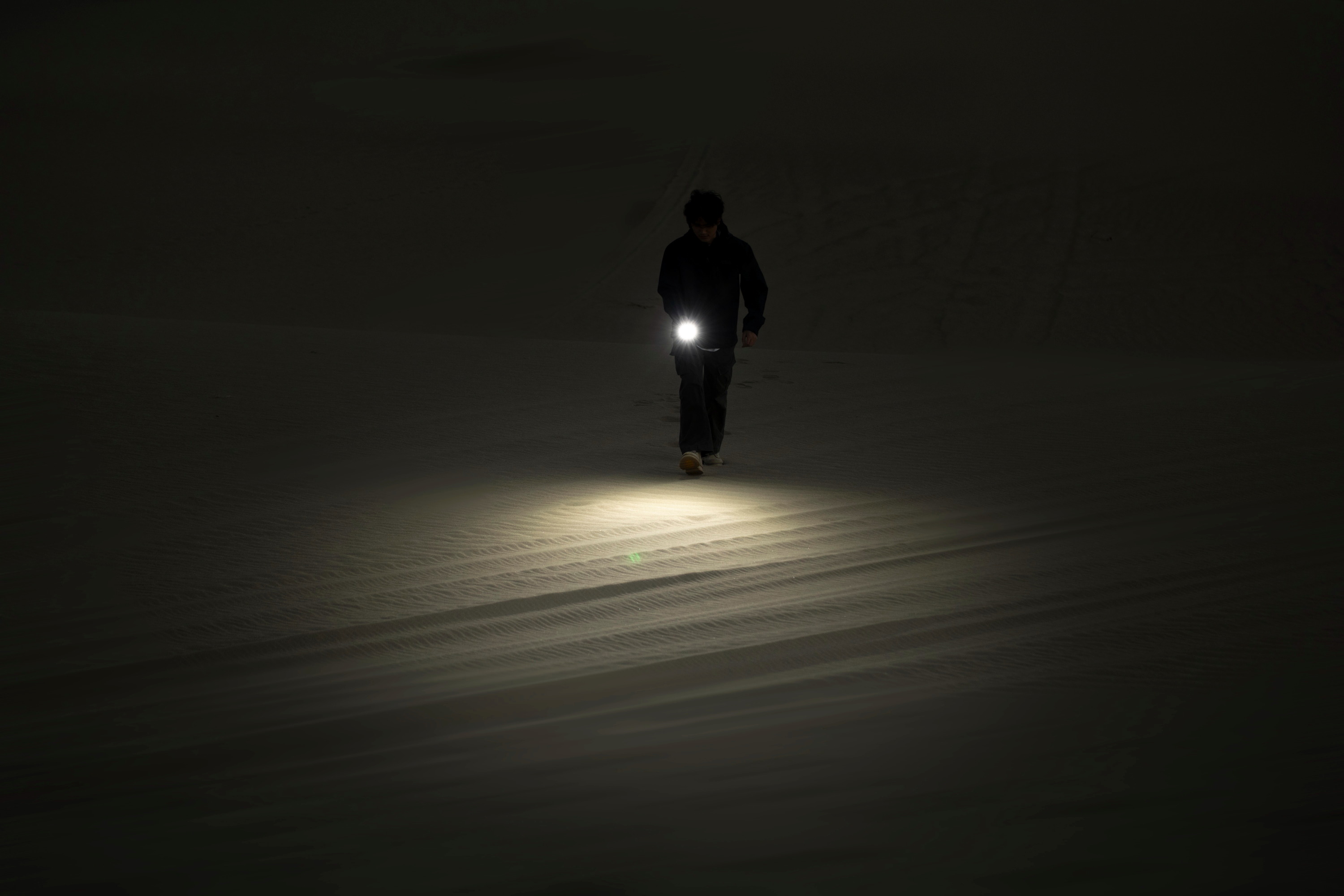 Persona caminando en la oscuridad de la noche por un terreno arenoso, usando una linterna para iluminar su camino.