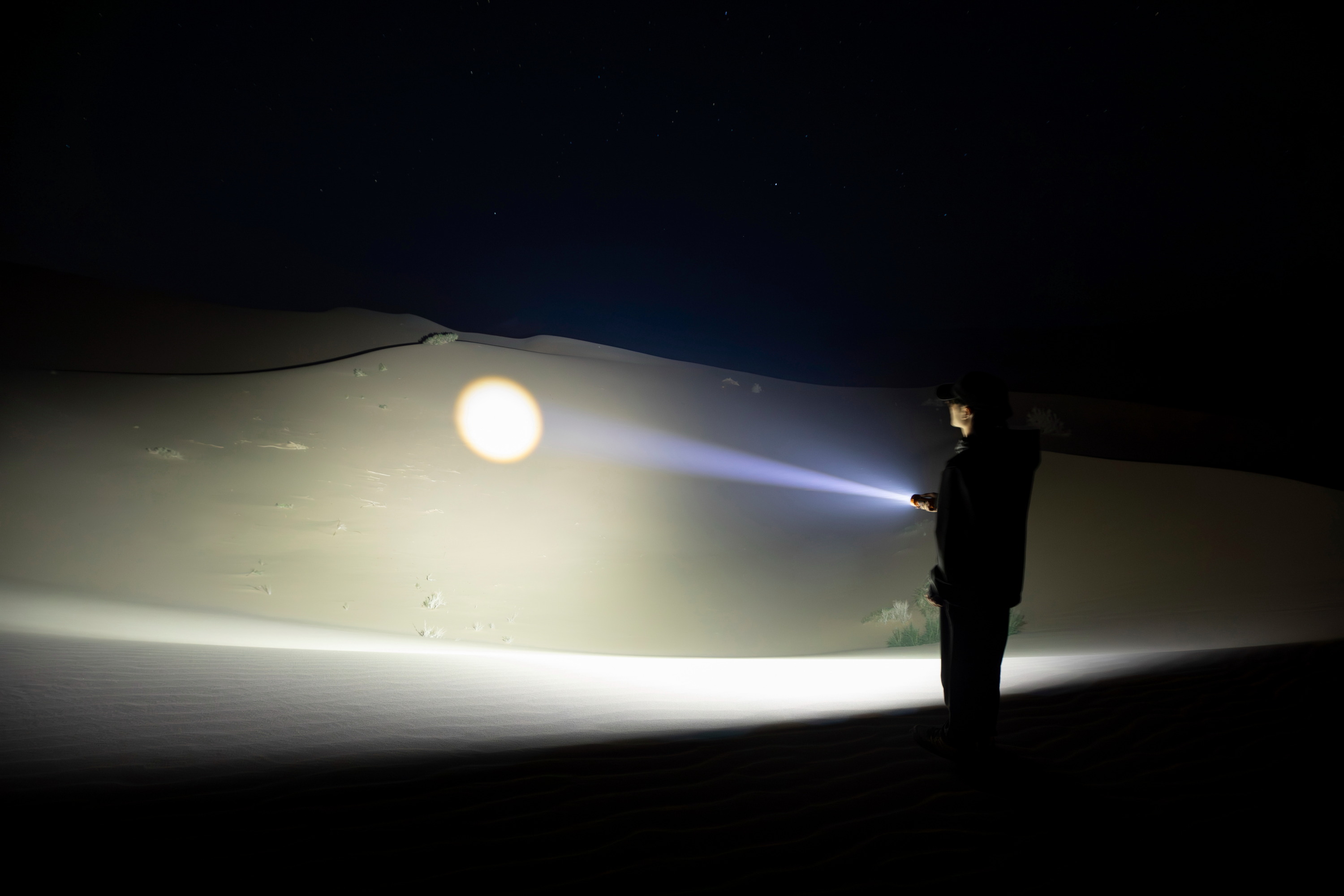 Explorador con linterna potente en dunas desérticas de noche, revelando el paisaje bajo el cielo estrellado.