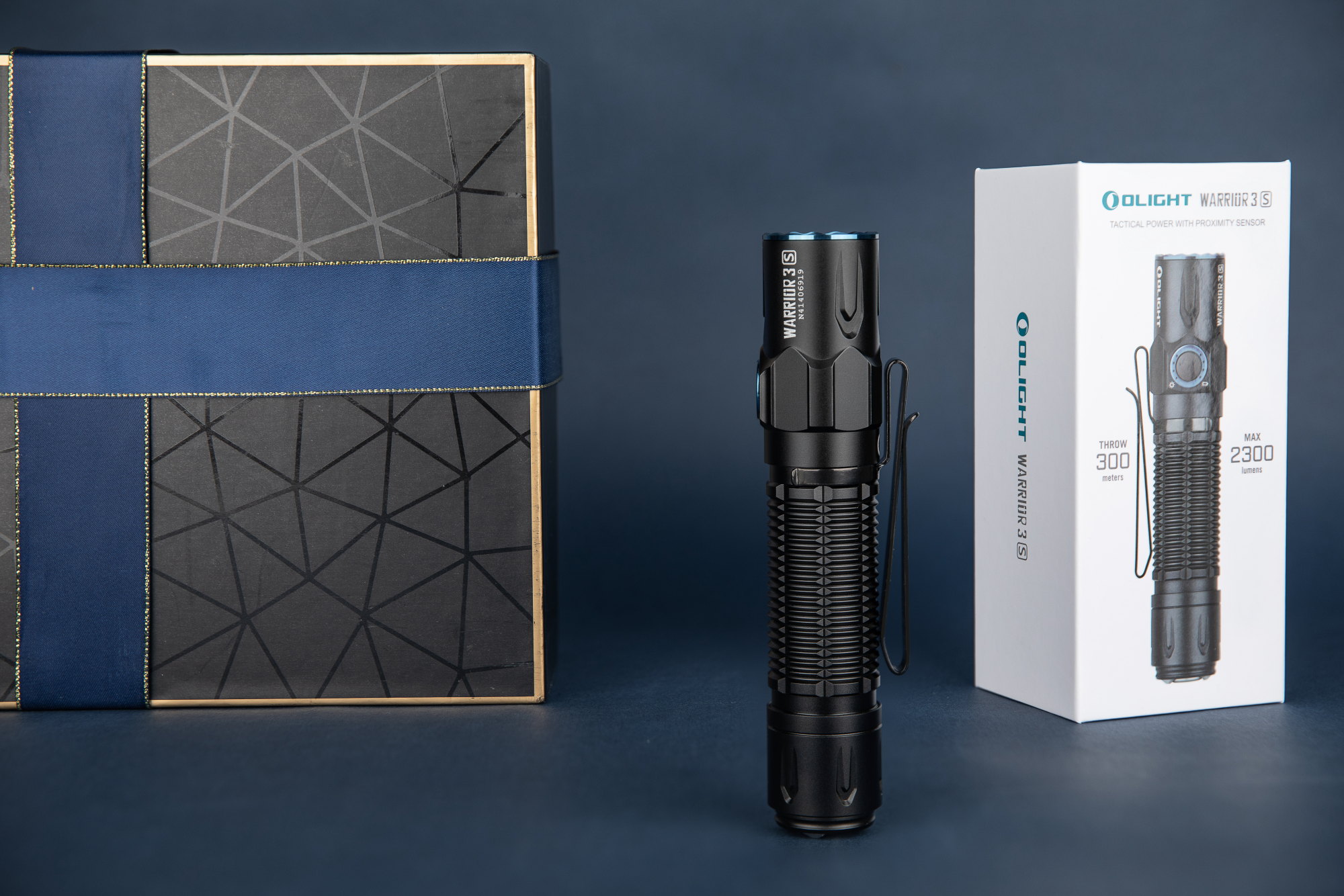 Lampe torche tactique Olight Warrior 3S (2300 lumens, 300m portée) avec capteur de proximité, présentée à côté de son emballage et d'un coffret cadeau bleu.