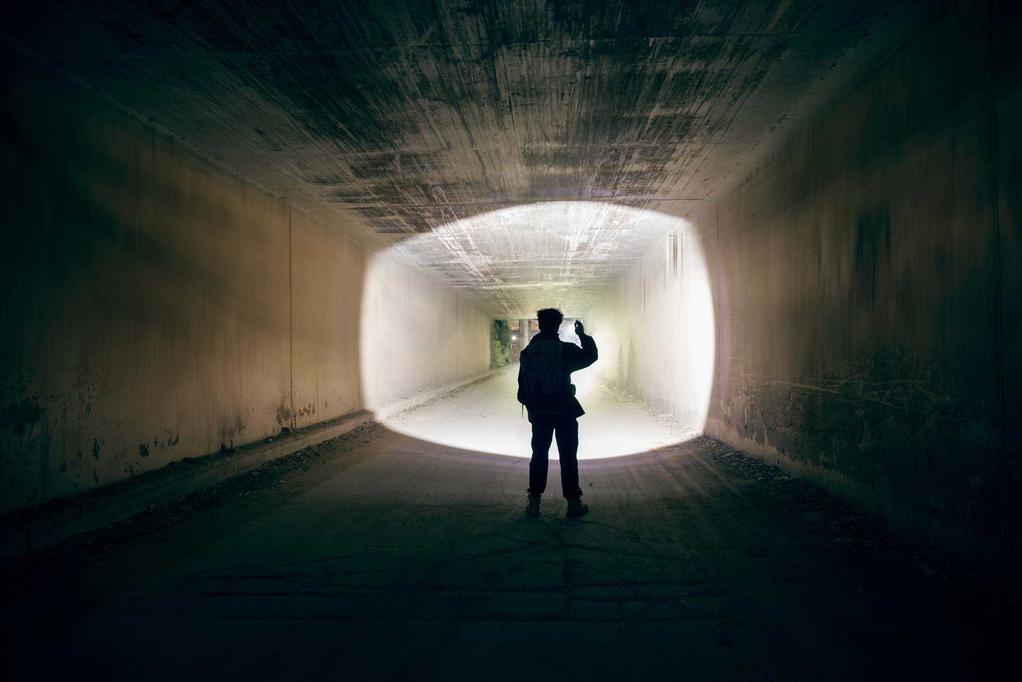 Hombre con linterna explorando un túnel oscuro, su silueta contra una luz brillante al final.