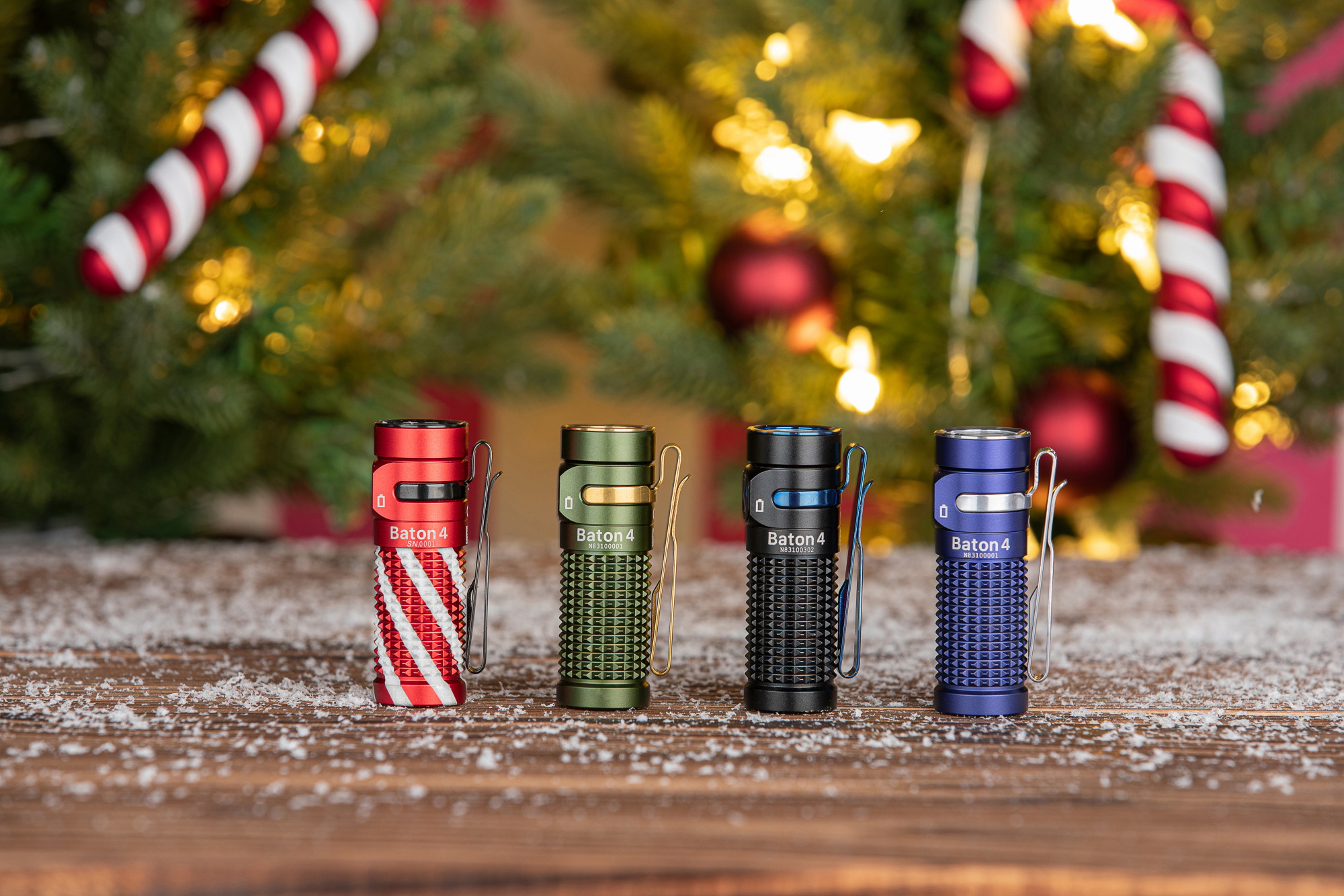크리스마스 장식 트리와 눈 덮인 테이블 위에 놓인 빨강, 초록, 검정, 파랑색 Baton 4 손전등.