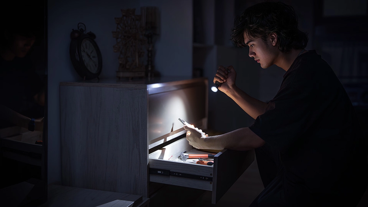 Man holding flashlight in dark room while searching through drawer with tools during power outage or emergency repair.