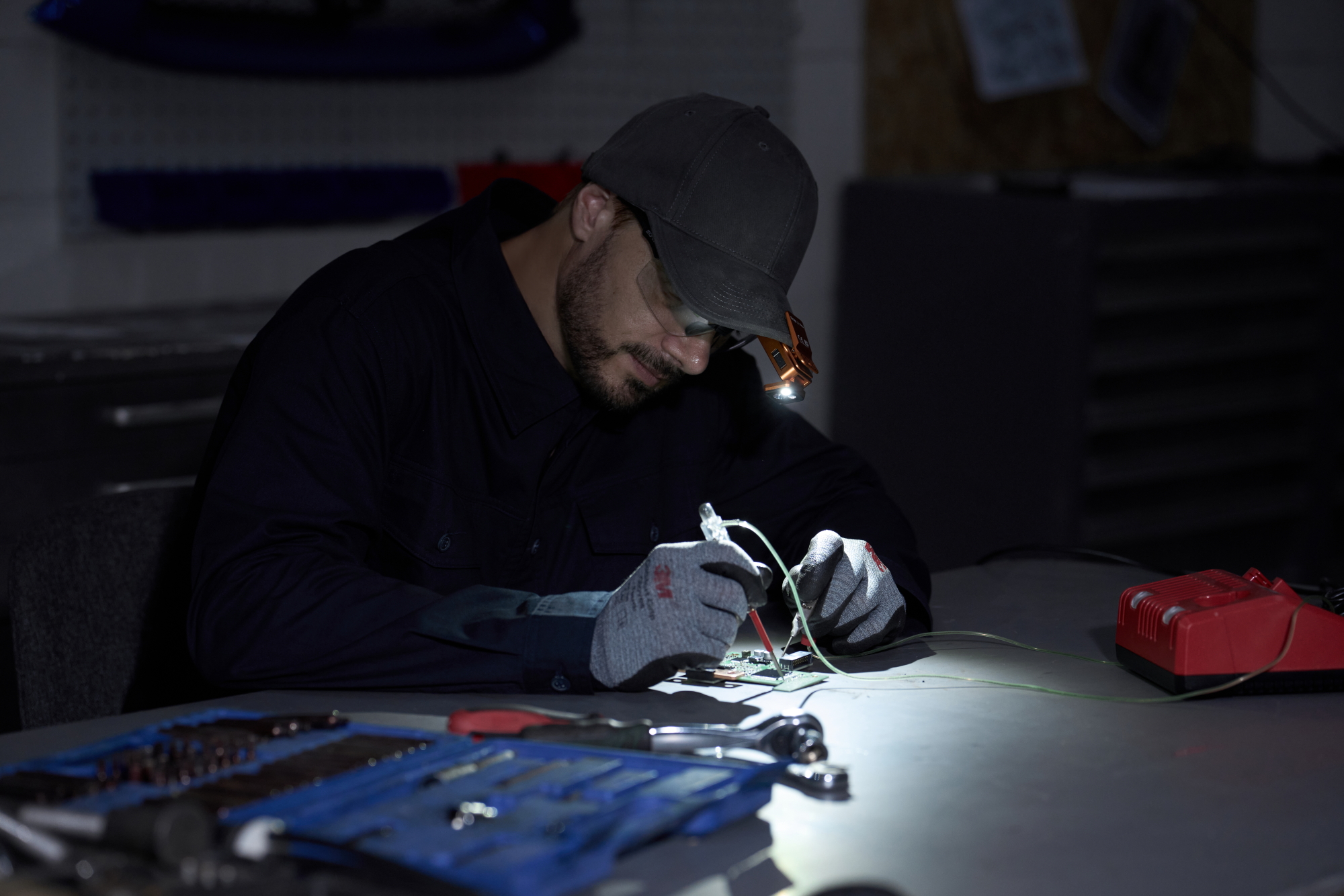 Technician wearing a cap-mounted LED light repairs an electronic circuit board at a dark workbench with tools, focusing on precision micro-soldering and hardware diagnostics.