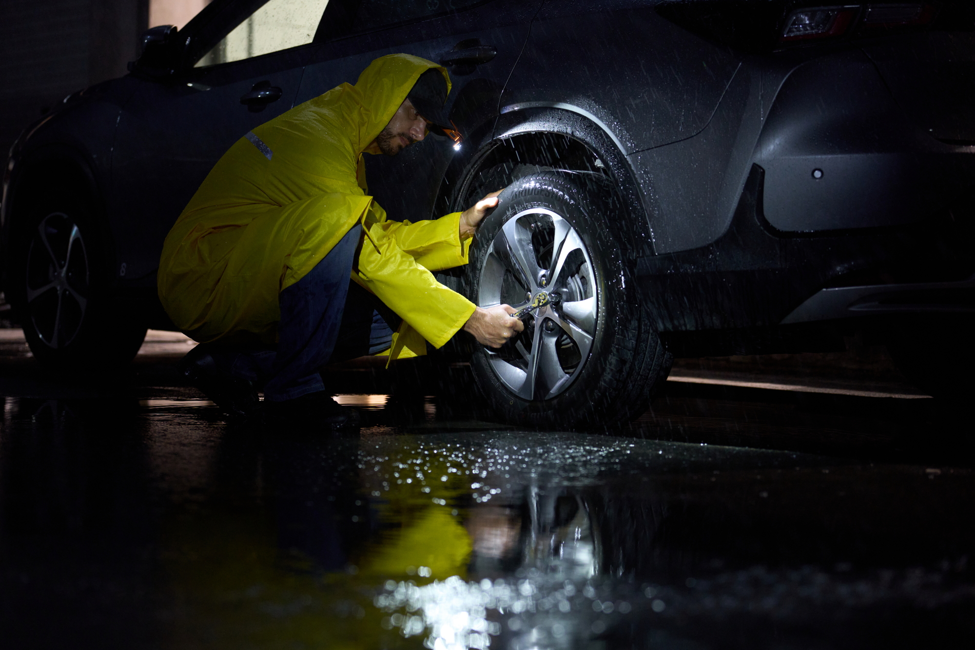 Man in a yellow raincoat changing a flat tire on a car at night in the rain, using a lug wrench and wearing a headlamp.