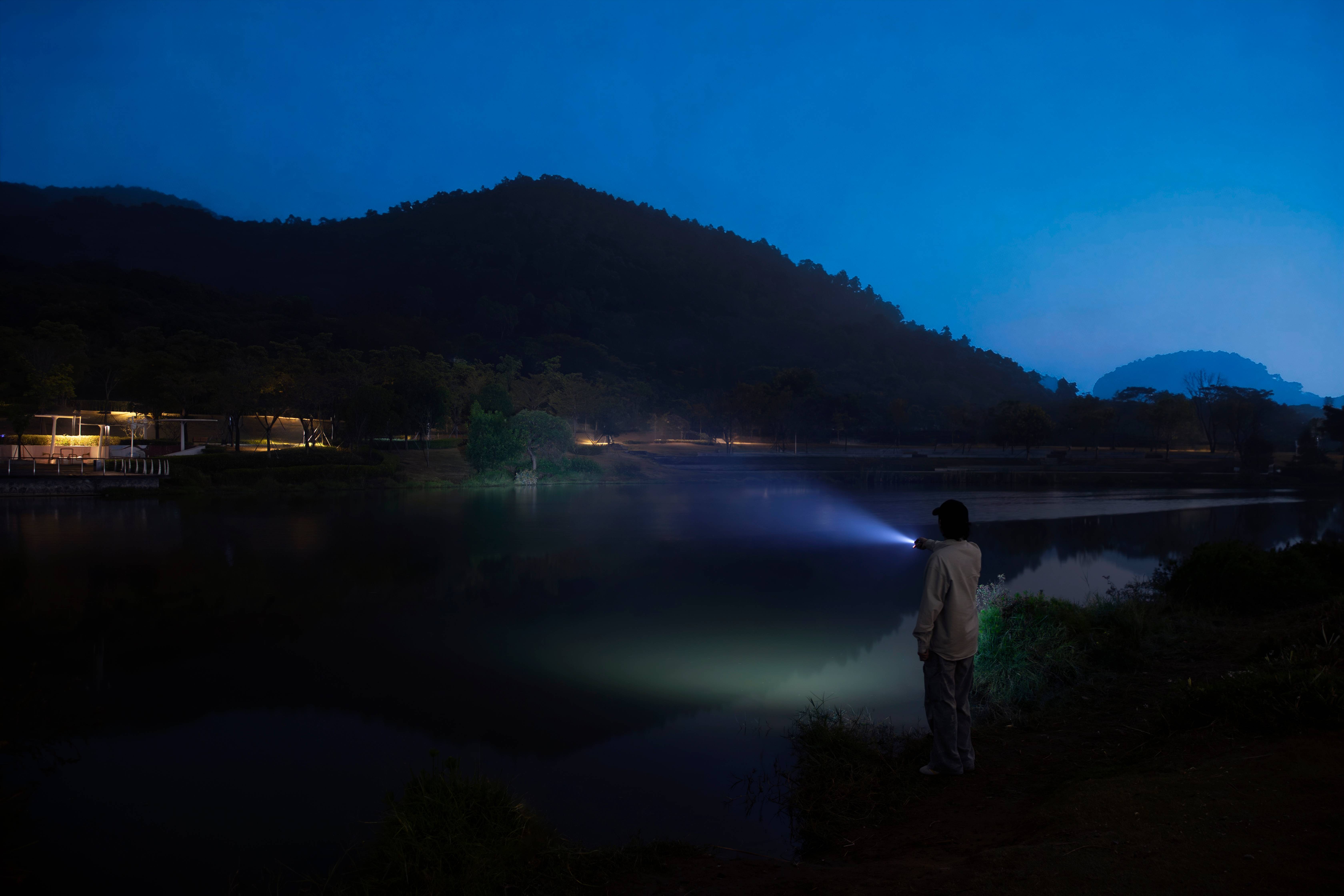 Persona utilizando una linterna LED potente de gran alcance frente a un lago y monta&ntilde;as durante la noche, equipo de iluminaci&oacute;n profesional para camping y actividades al aire libre.