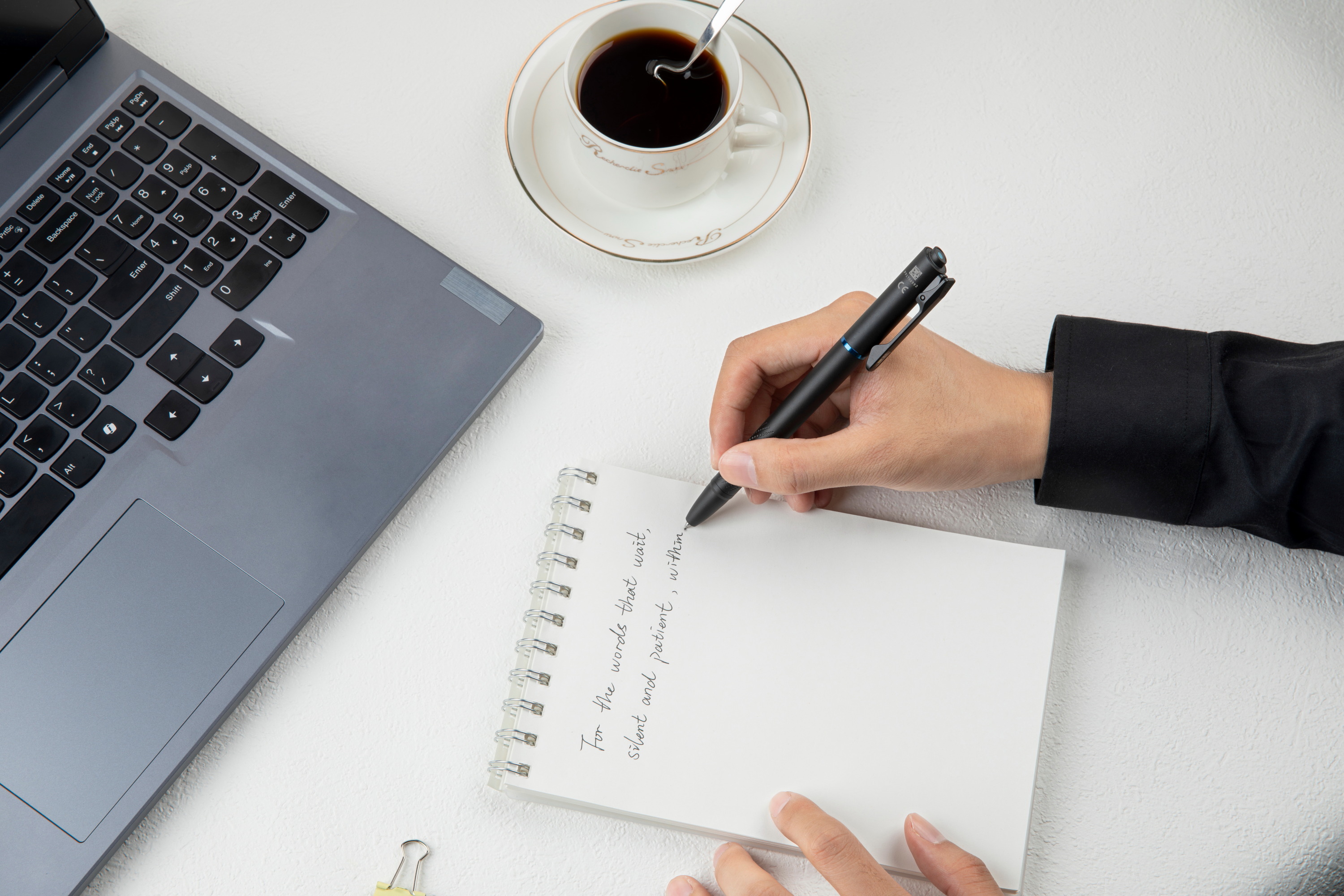 Person writing in a spiral notebook on a white desk with a laptop and a cup of black coffee. Professional workspace for creative writing, journaling, and productivity.