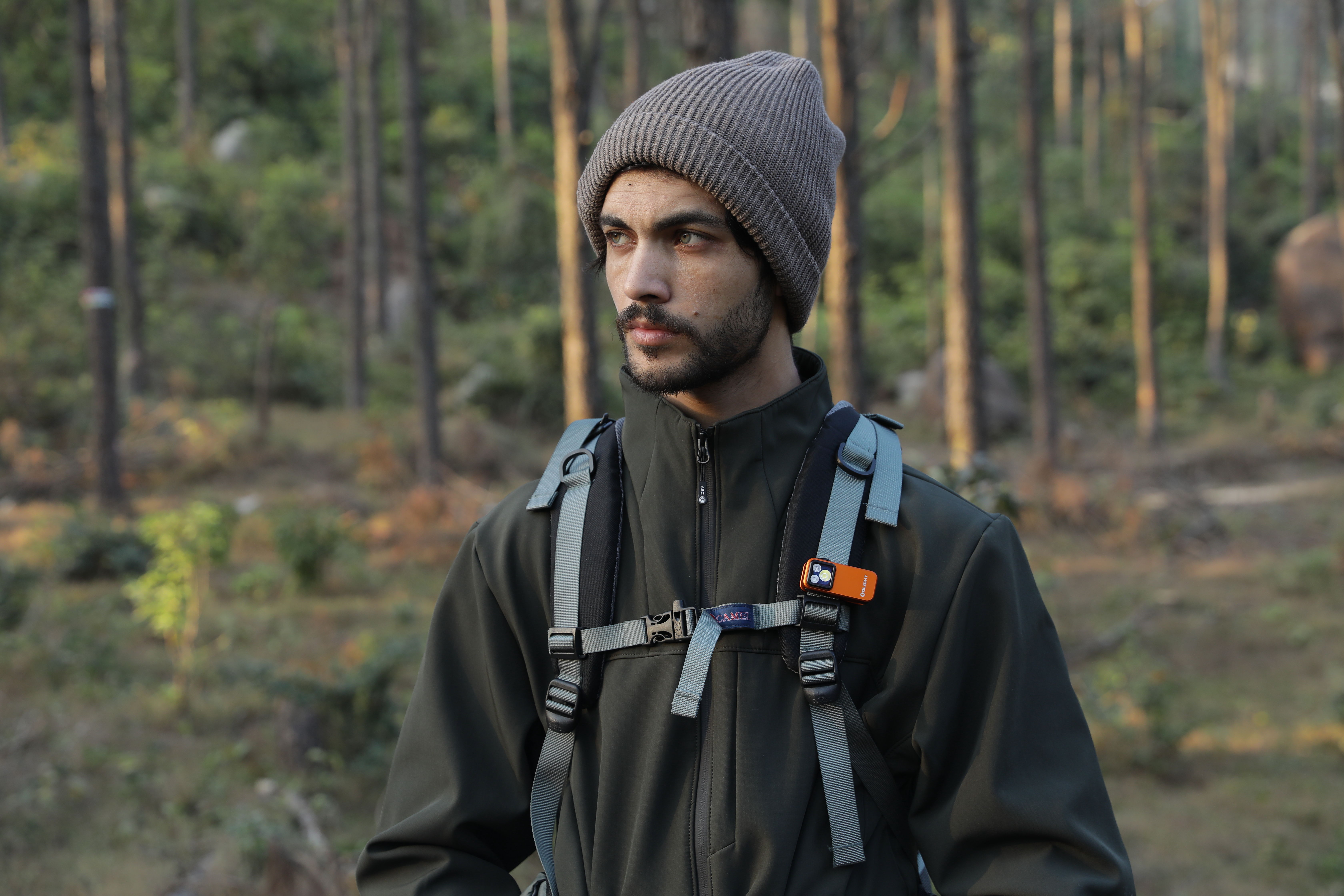 Outdoor enthusiast hiking in a forest with a portable orange clip-on EDC flashlight attached to his backpack strap.