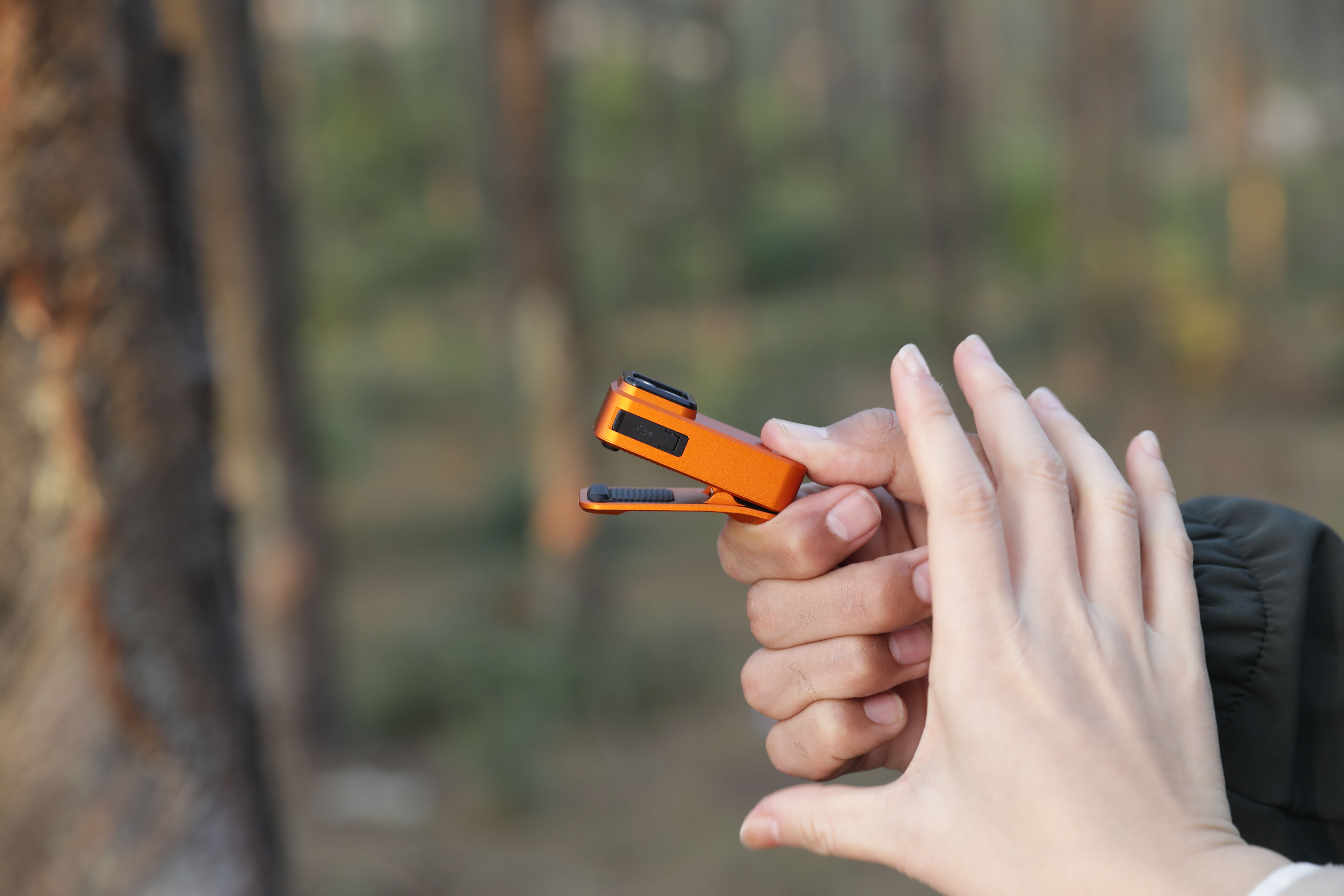Person holding a compact orange fingertip pulse oximeter in a forest, monitoring blood oxygen levels and heart rate during outdoor activities like hiking.