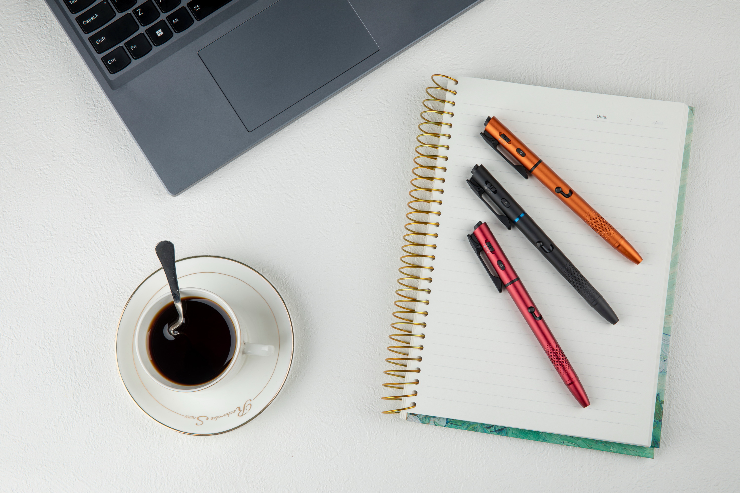Espace de travail avec ordinateur portable, tasse de caf&eacute; noir et carnet de notes &agrave; spirale avec stylos color&eacute;s sur un bureau blanc.