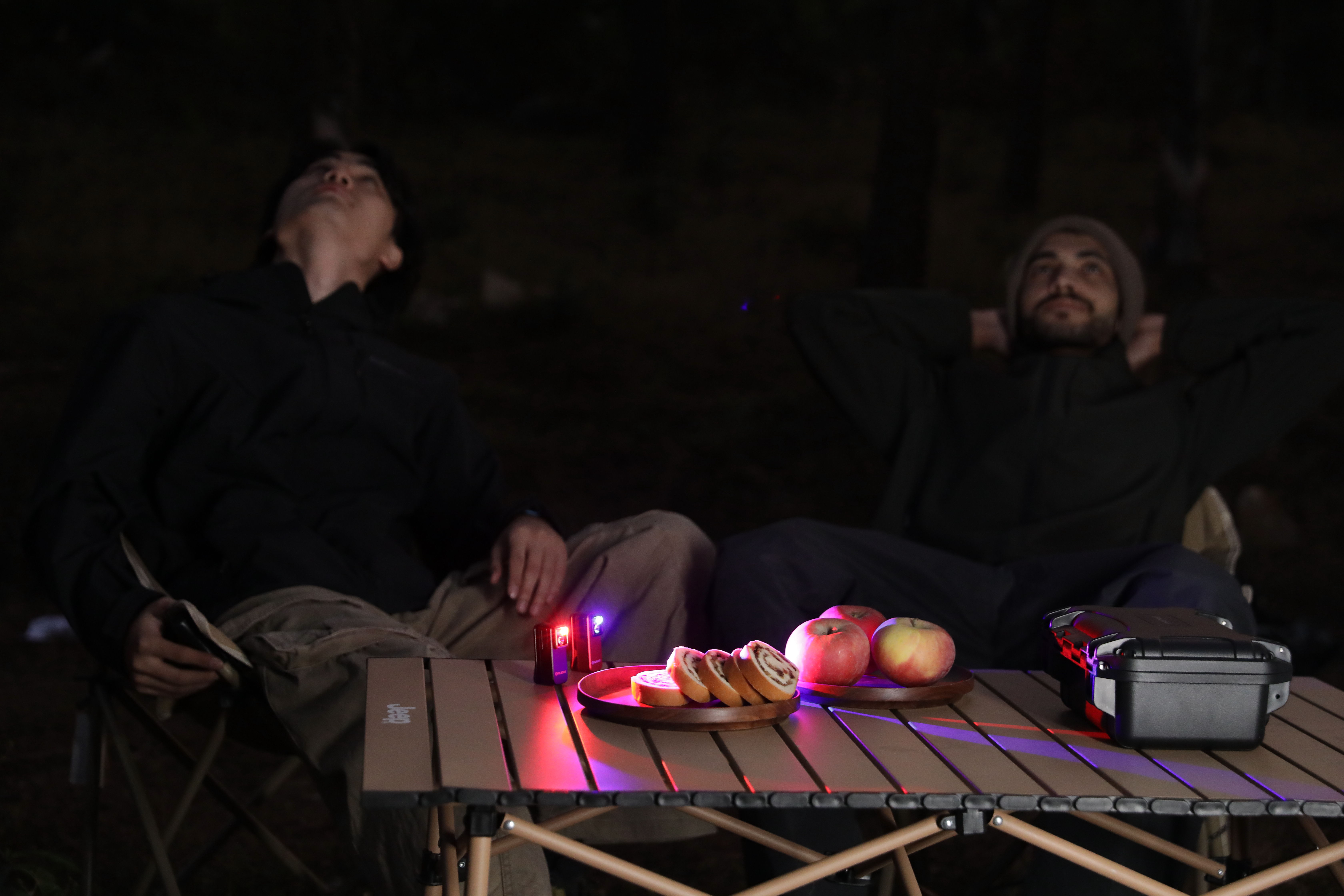 Two men relaxing at a night campsite behind a folding table with Olight Oclip small clip-on lights providing signal lighting.
