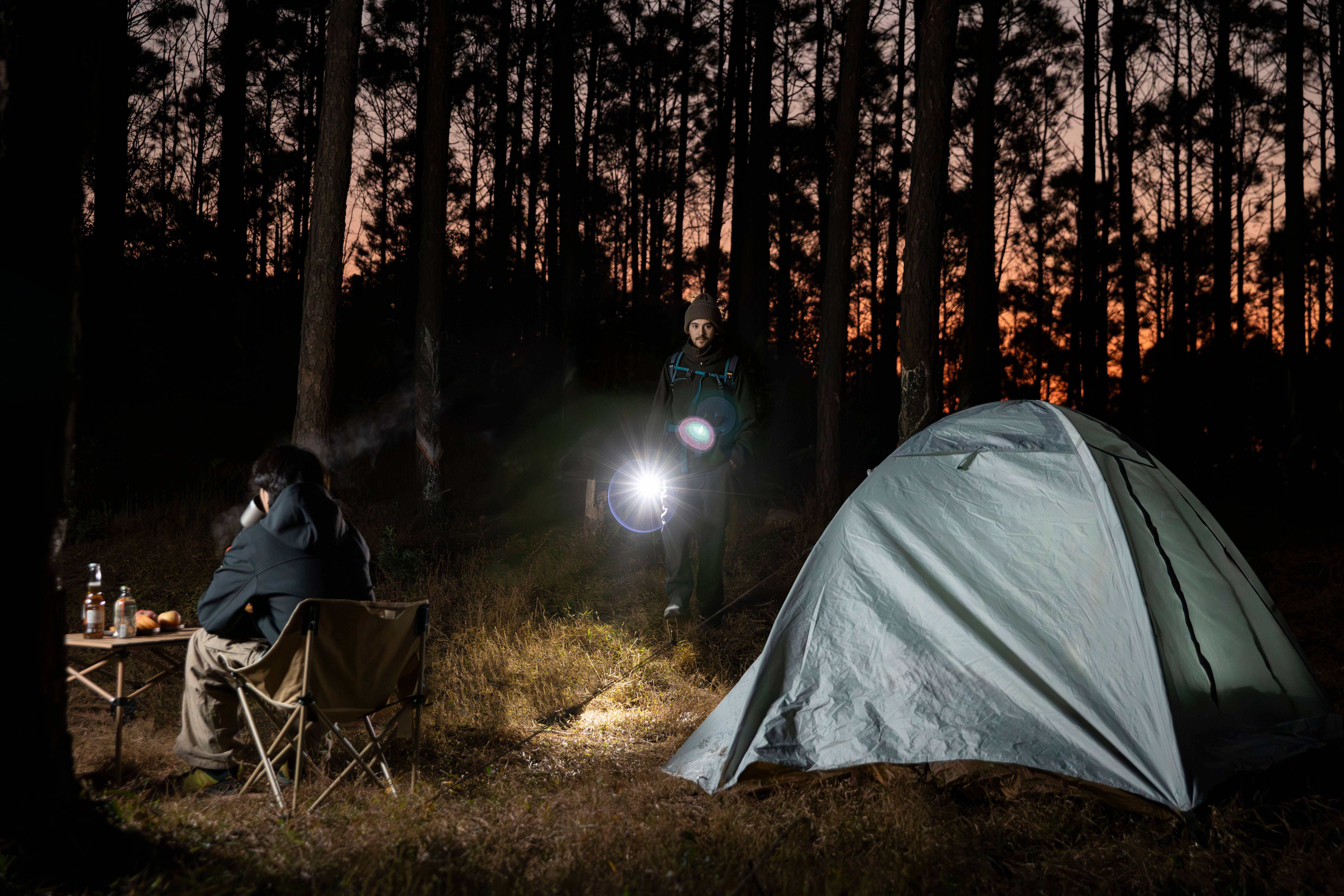 A hiker using a powerful flashlight at a night campsite in a pine forest, featuring a grey camping tent and a person sitting by a folding table at dusk.