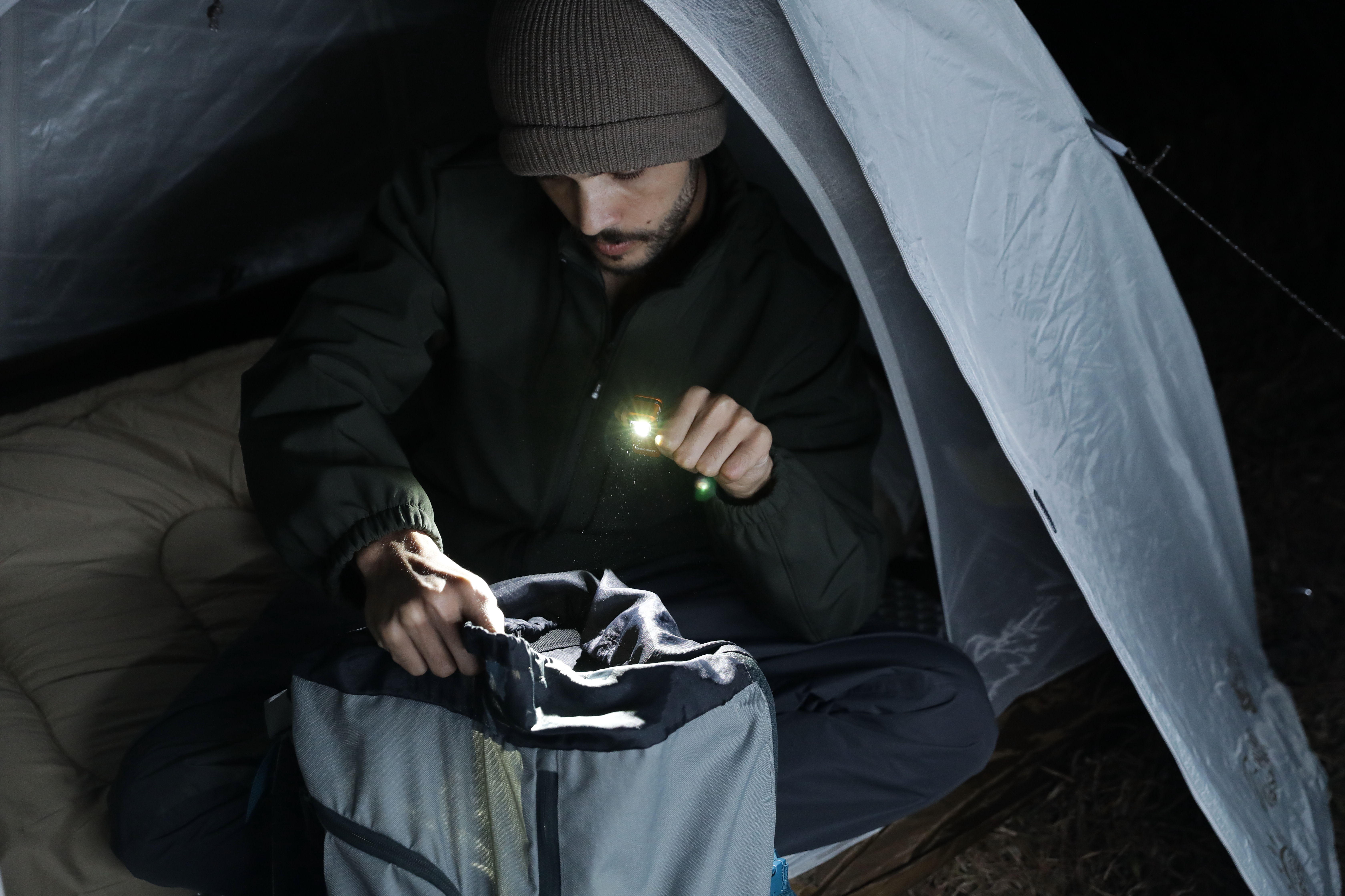 Man using a compact LED flashlight to search through a backpack inside a tent while camping at night.