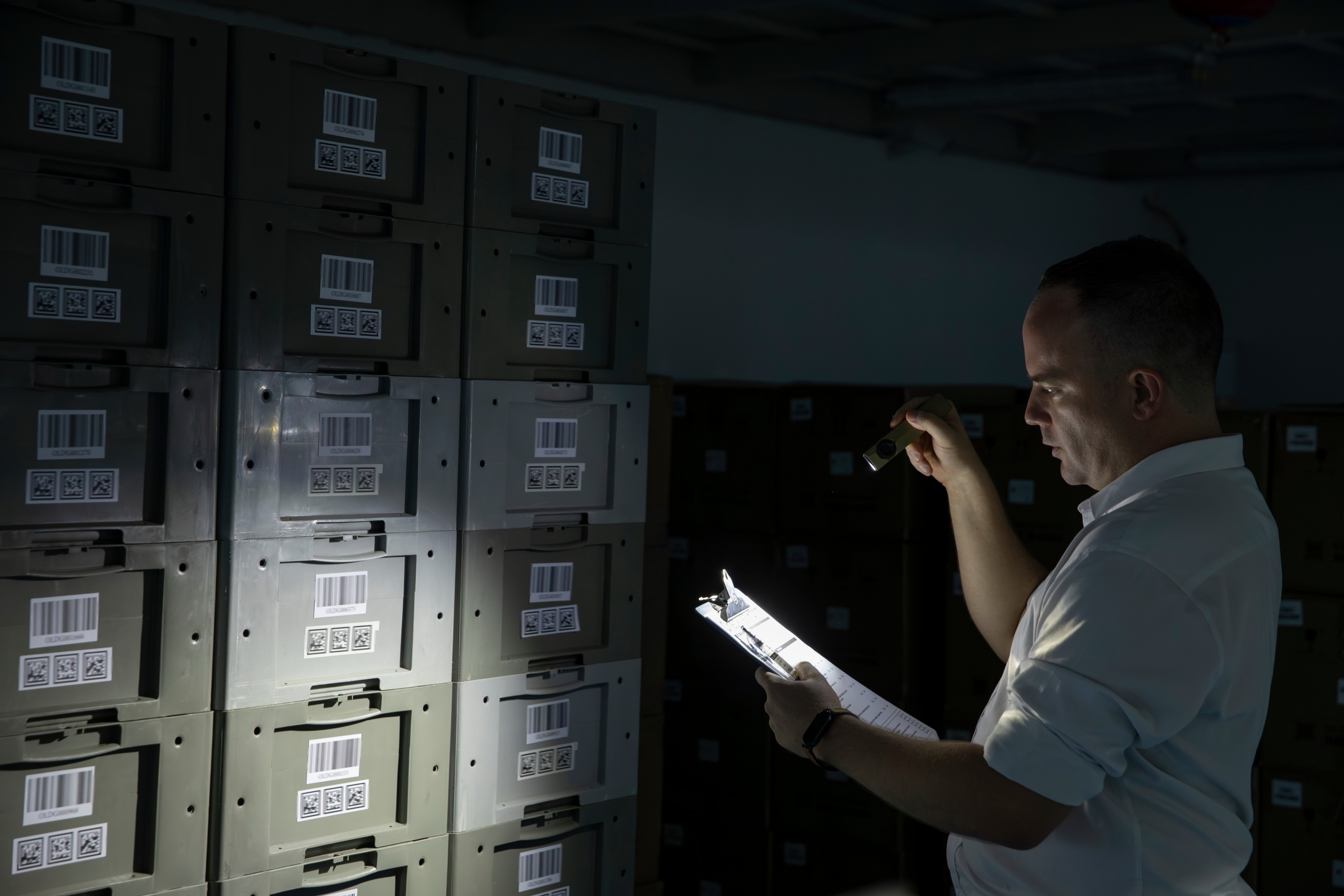 Warehouse worker using a flashlight and clipboard to conduct an inventory audit of stacked barcoded storage bins in a dark logistics facility.