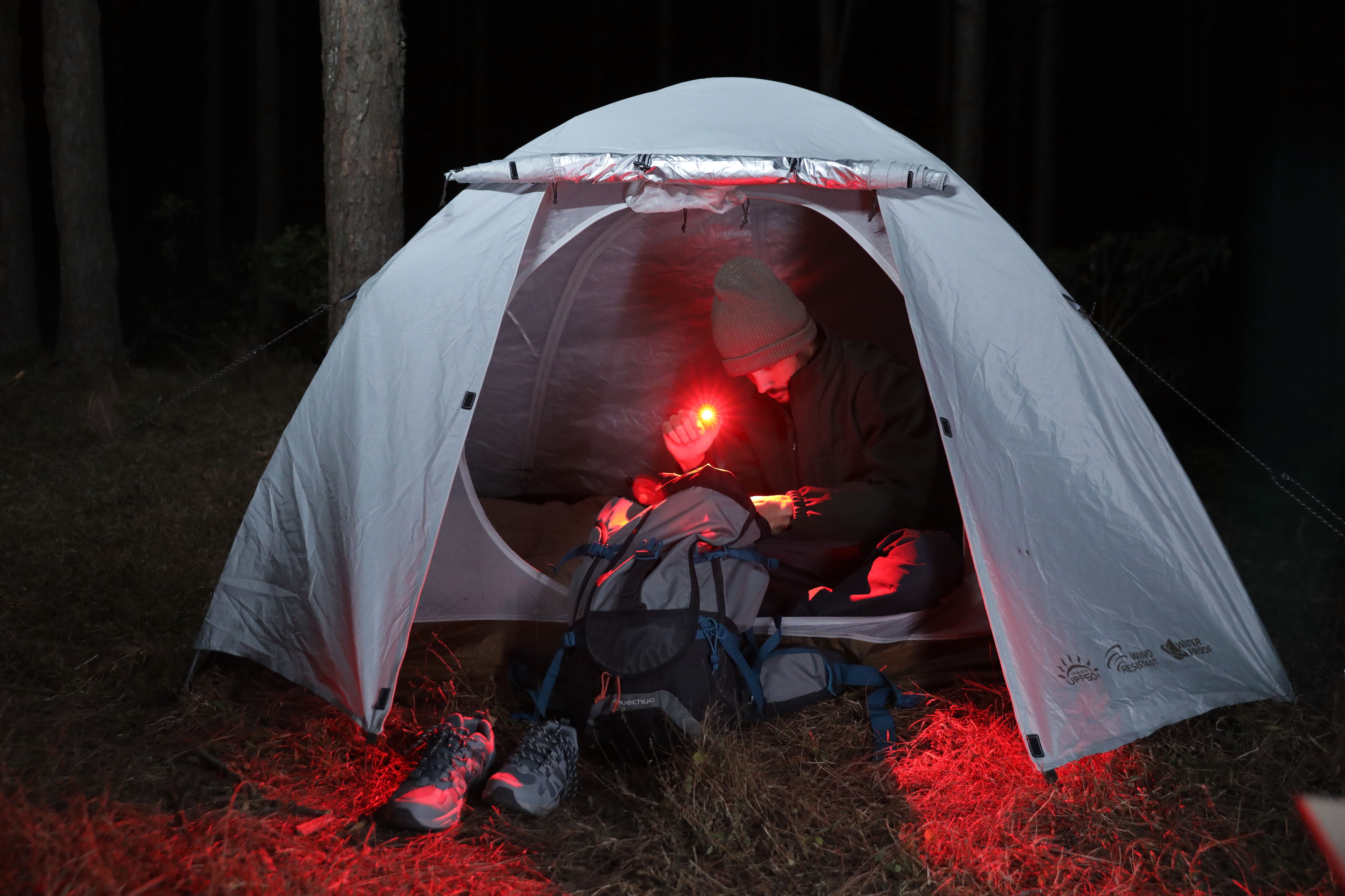 Man using a red light to search through a backpack inside a white camping tent at night in a forest, with hiking boots placed outside.