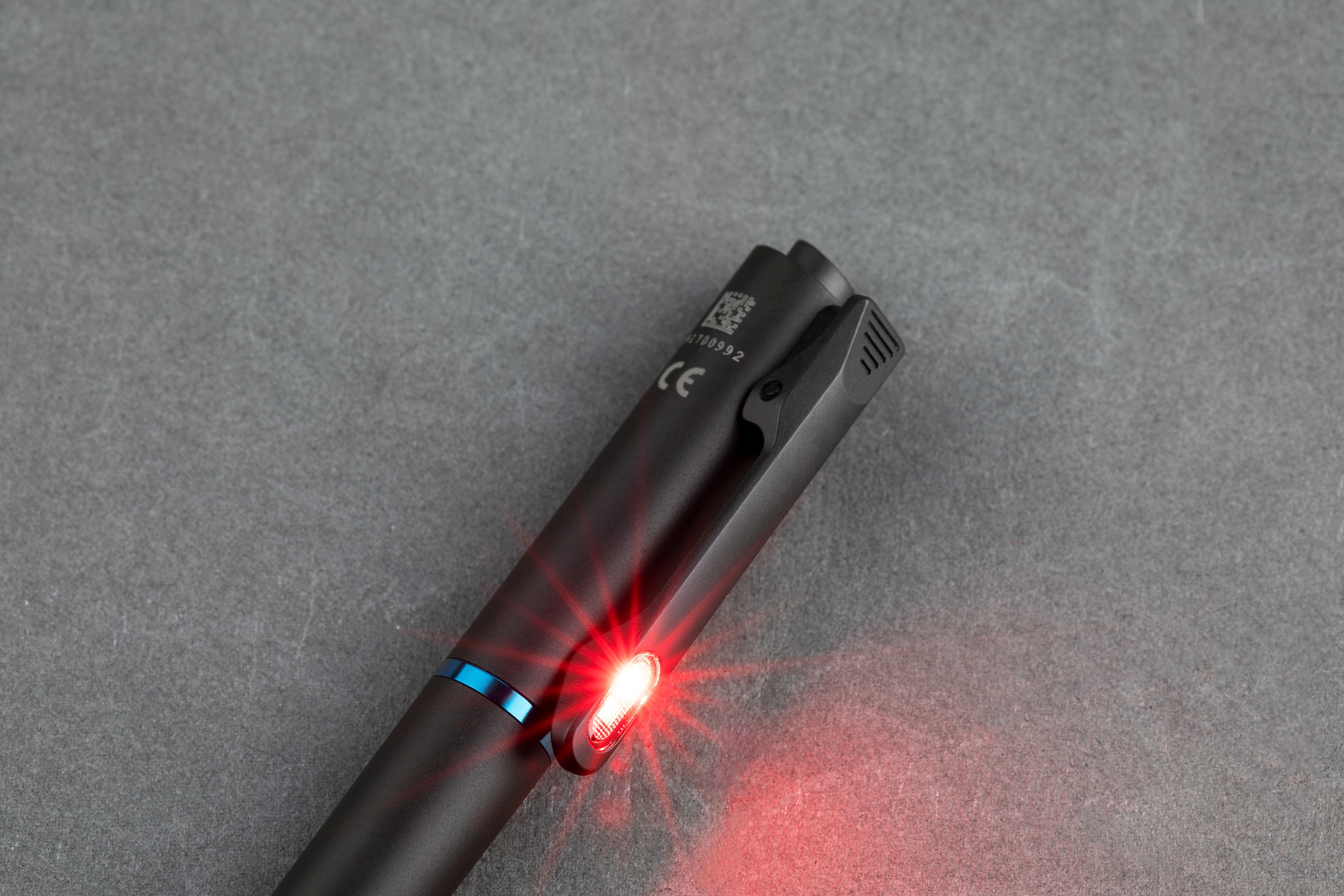 Black EDC penlight with an active red LED light on the pocket clip, placed on a gray textured background, highlighting its tactical and low-light utility features.