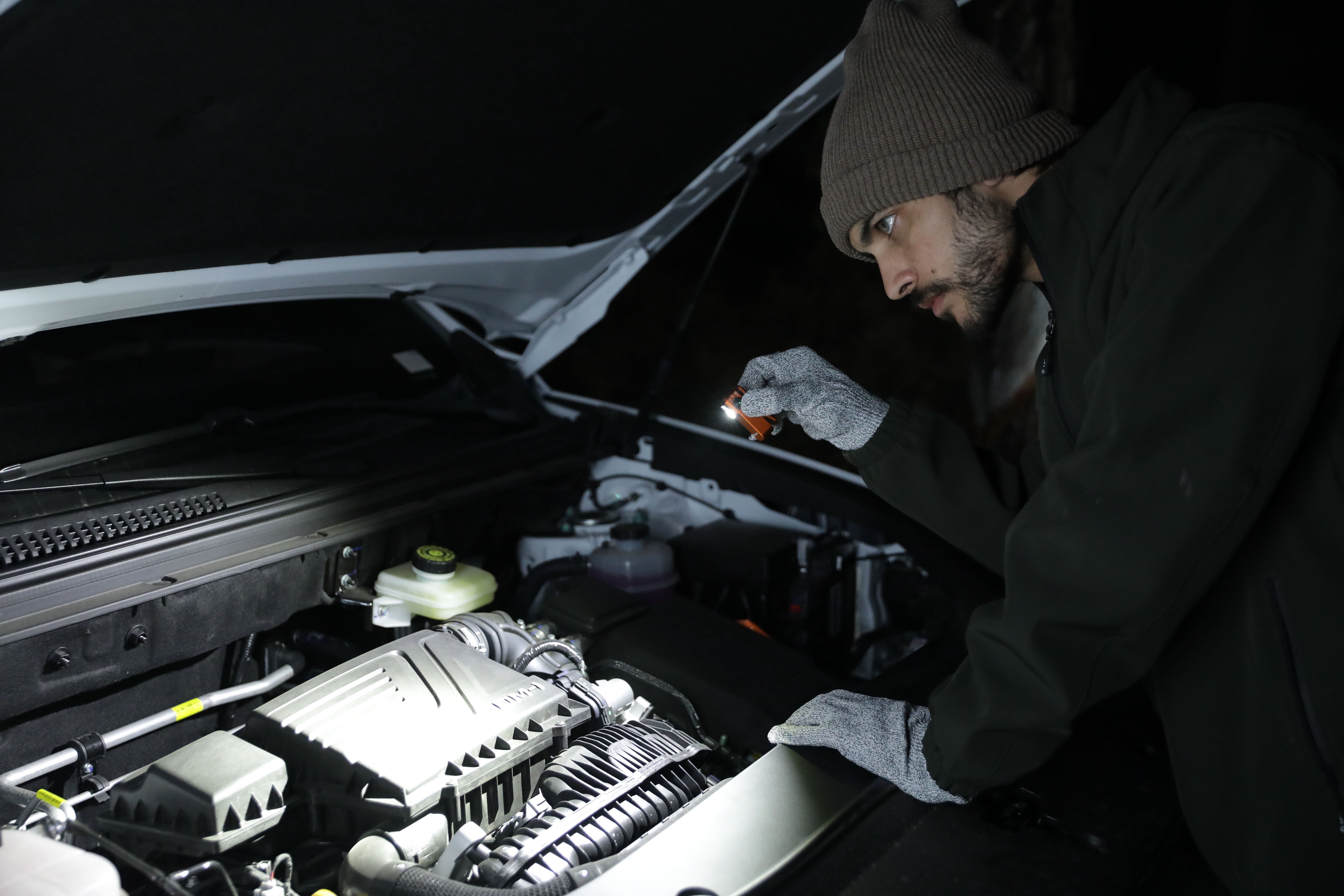 Man in beanie and gloves using a flashlight to inspect a car engine under the hood at night.