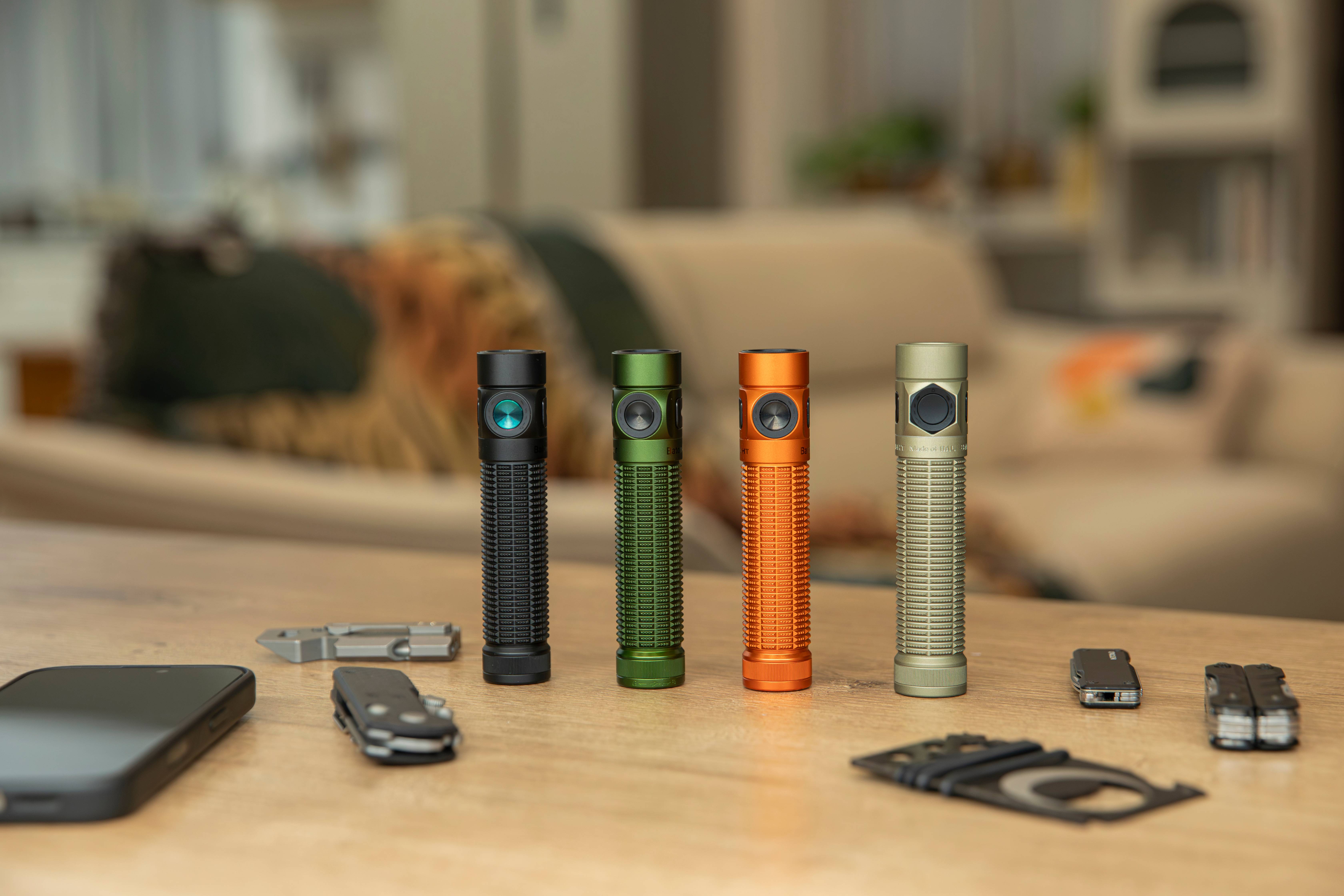 Collection of compact LED torches in black, green, orange, and tan, displayed on a wooden table alongside EDC gear.