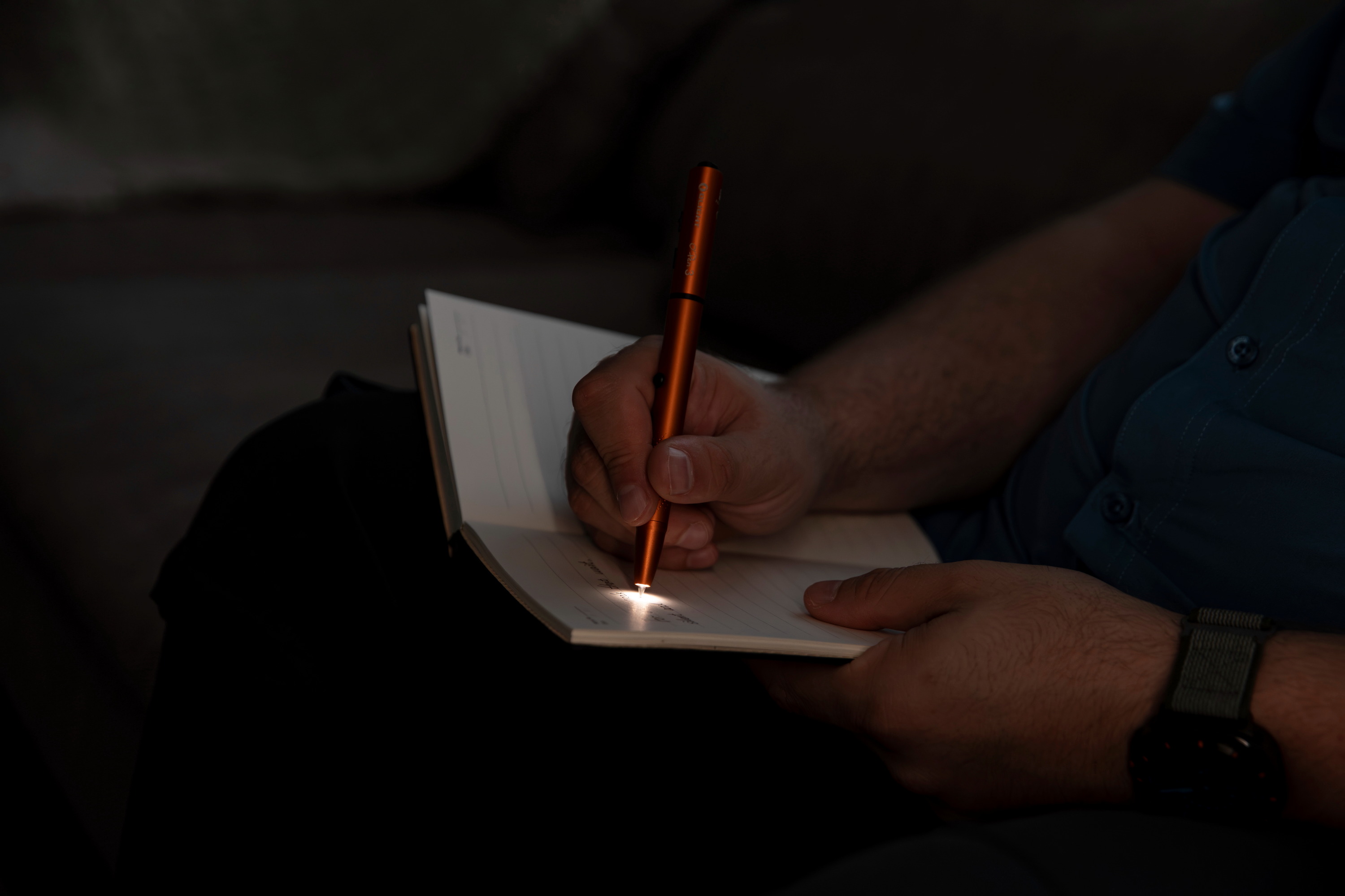 Persona utilizando un bol&iacute;grafo con luz LED naranja para escribir en un cuaderno en la oscuridad, destacando la funcionalidad de iluminaci&oacute;n t&aacute;ctica para tomar notas.