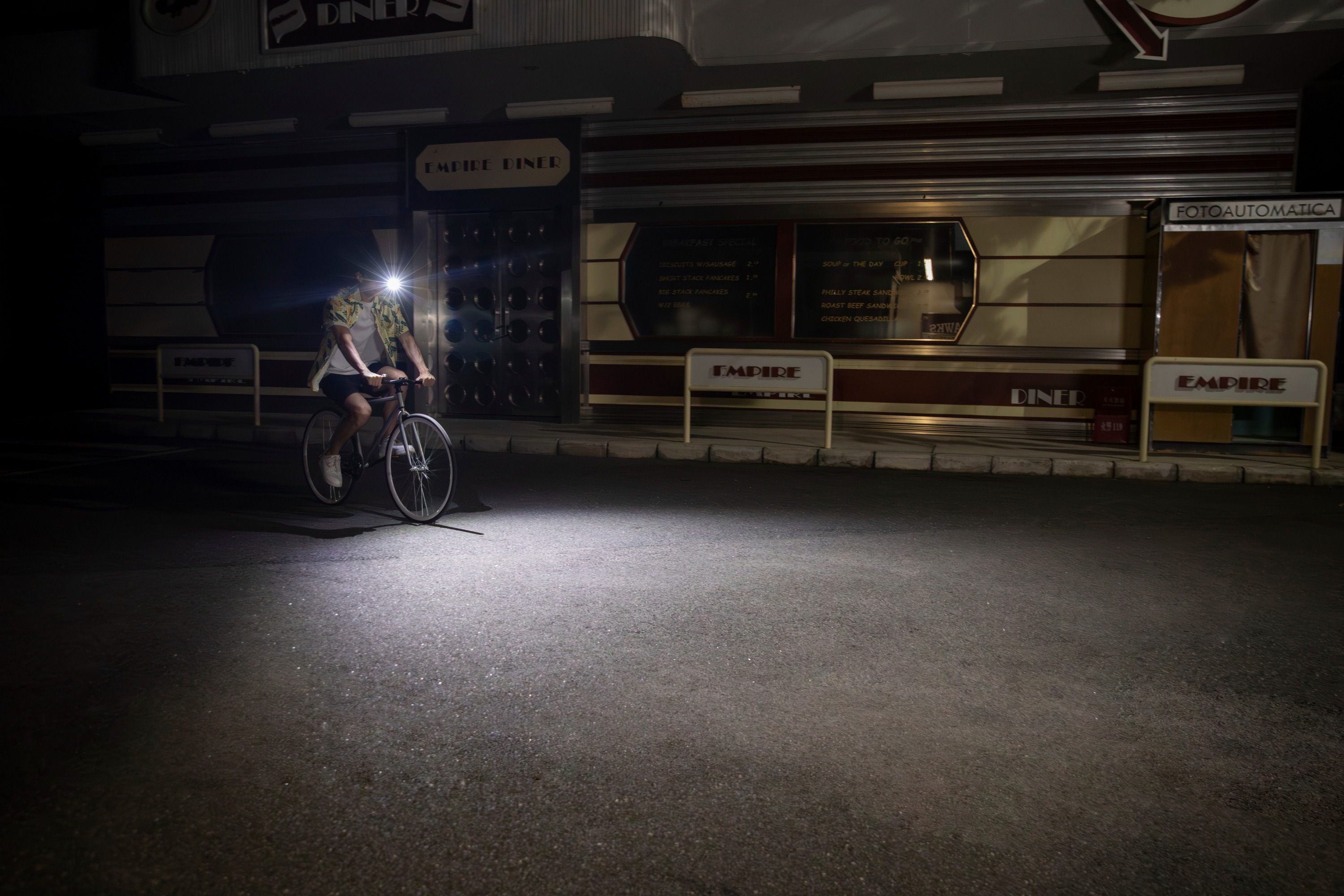 A cyclist riding a bicycle at night past the retro Empire Diner, illuminated by a bright headlight beam on the dark asphalt road.