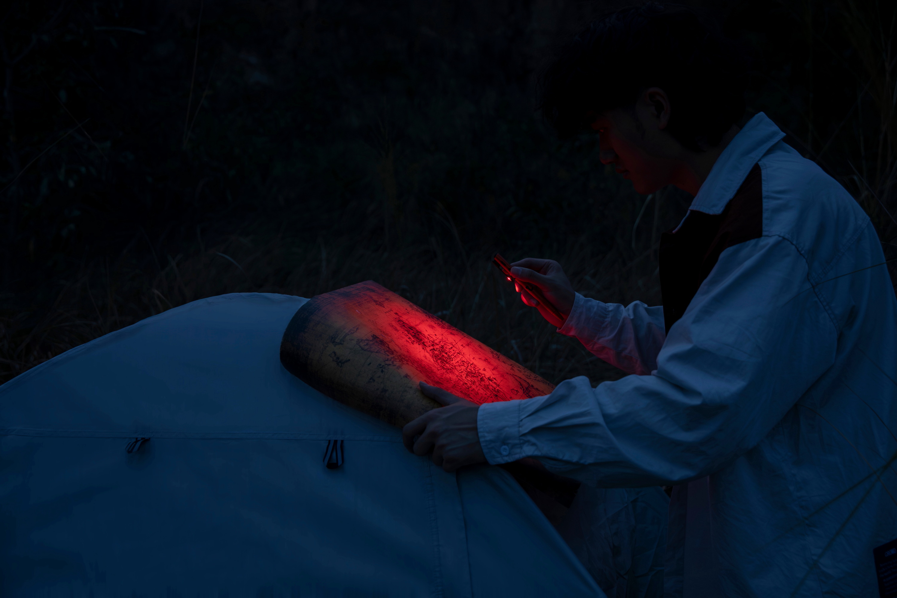 Explorador en un campamento nocturno utilizando una luz roja t&aacute;ctica para consultar un mapa sobre su tienda de campa&ntilde;a, preservando la visi&oacute;n nocturna durante una aventura al aire libre.