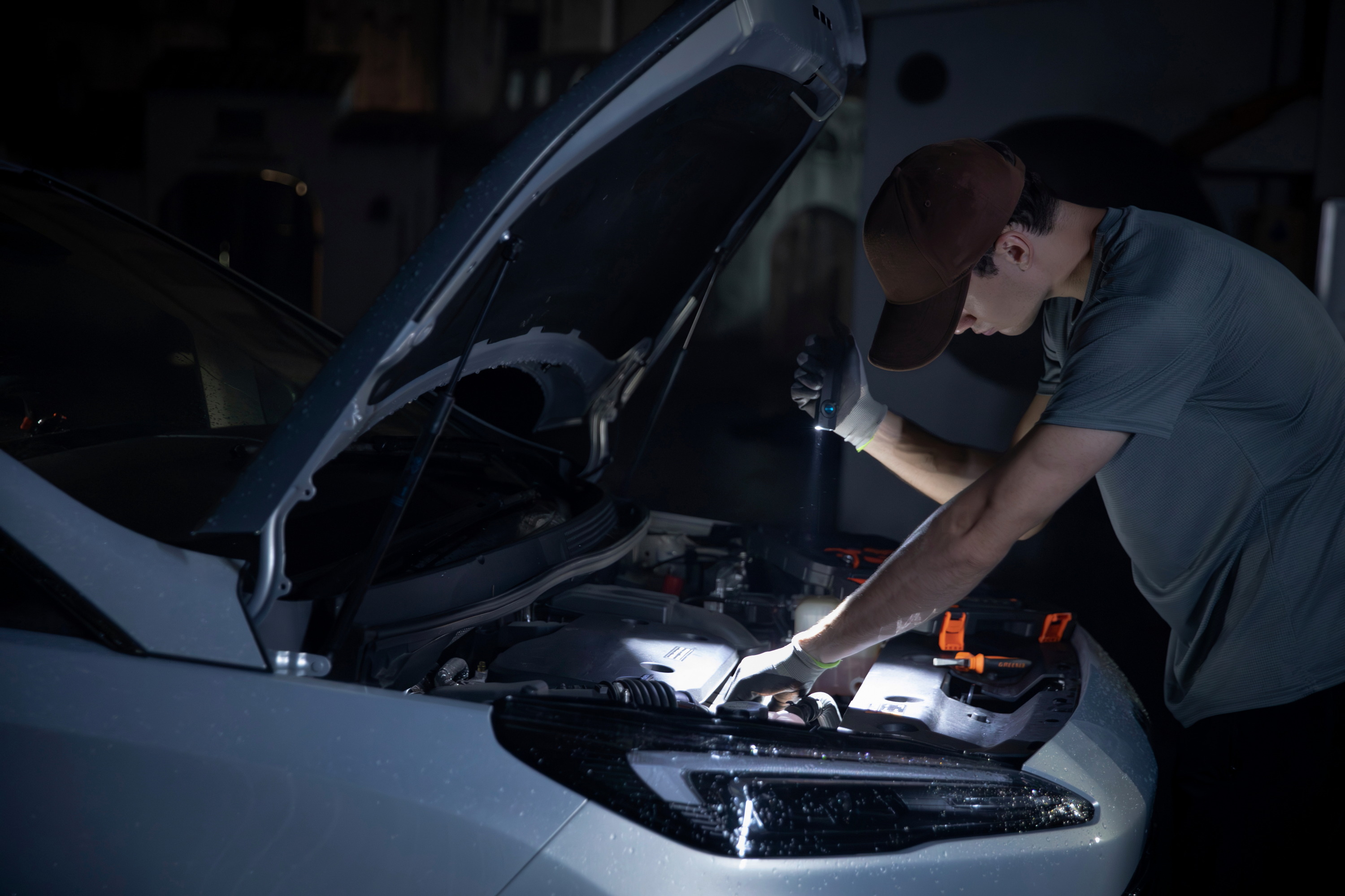 Mechanic with a flashlight inspects a car's engine bay at night. Automotive repair, vehicle maintenance, diagnostic service.