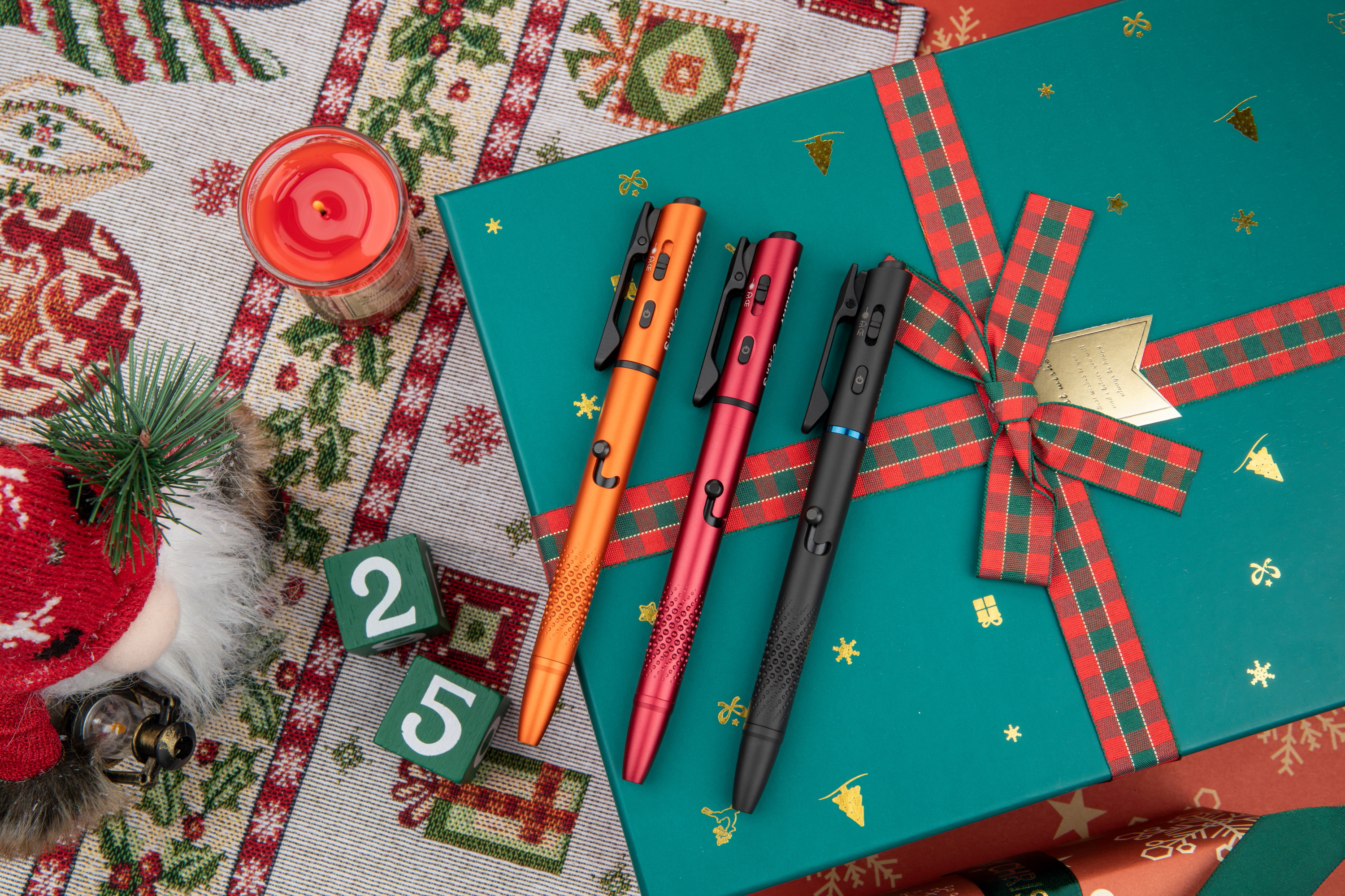 Olight multifunction pens (orange, red, black) on a festive teal Christmas gift box with plaid ribbon, surrounded by holiday decor, a lit candle, and '25' blocks.