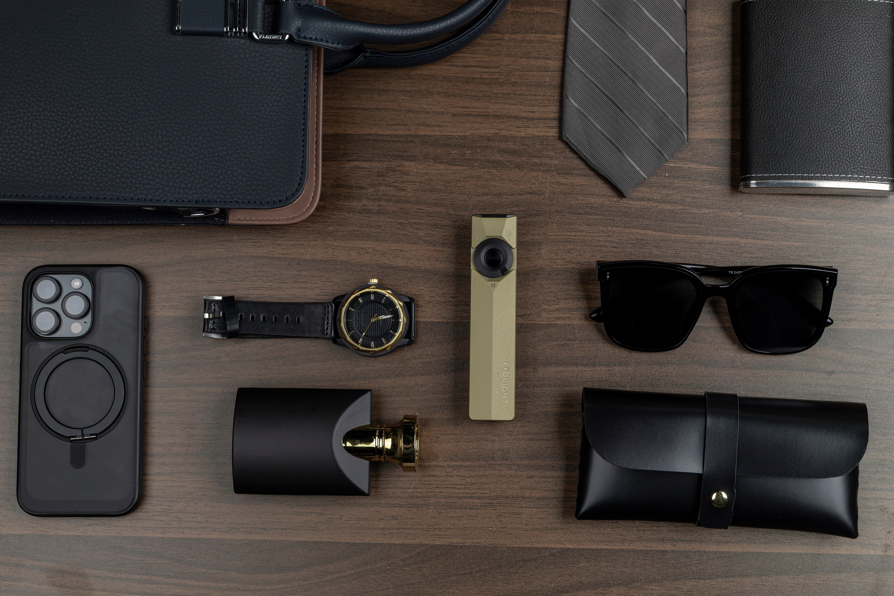 Premium men's everyday carry essentials flat lay featuring an Olight Arkfeld flat flashlight, smartphone, luxury watch, sunglasses, and leather accessories on a dark wooden background.