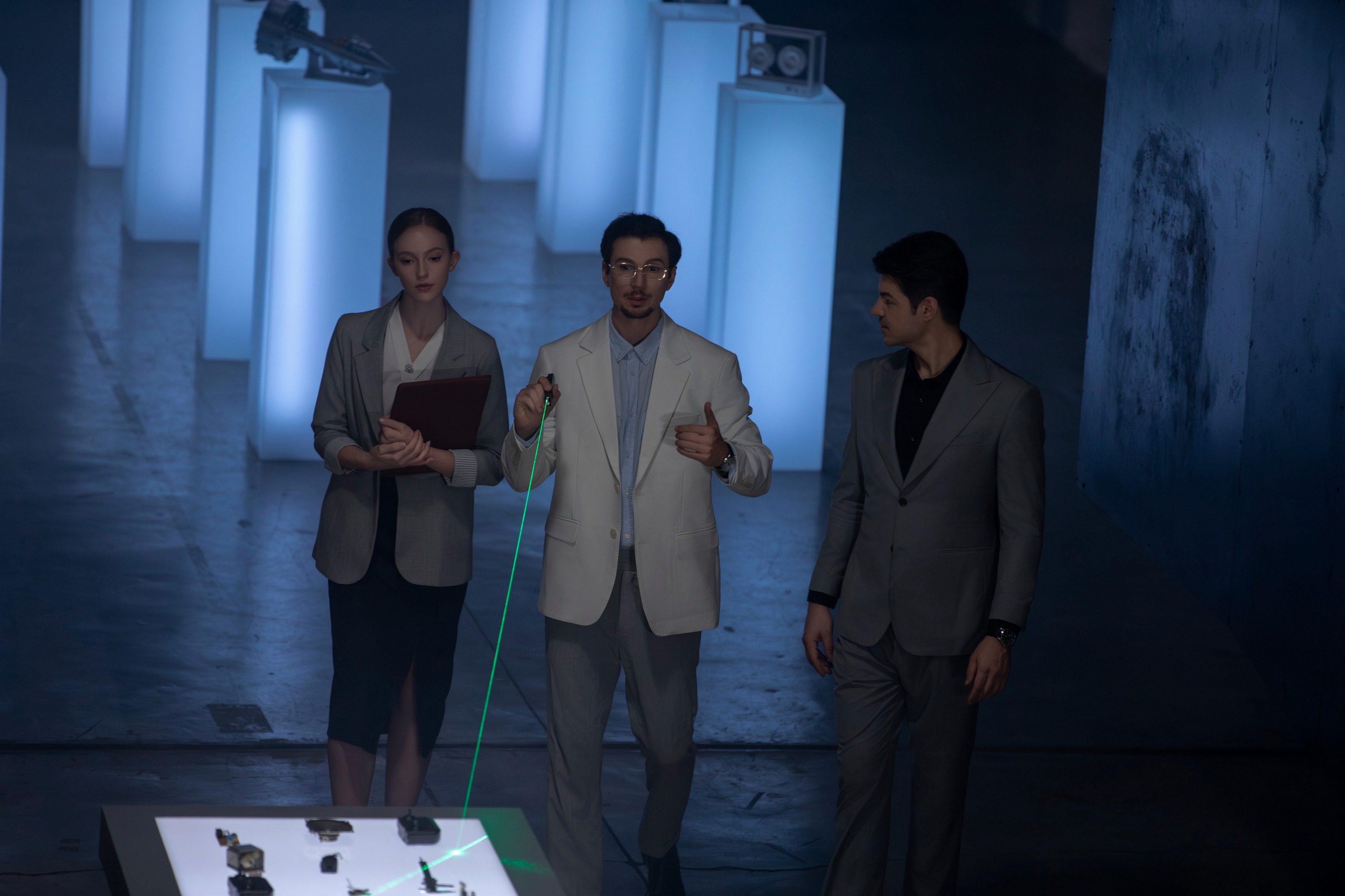 Professionals exploring a futuristic tech exhibition. A man uses a green laser pointer to demonstrate artifacts on a glowing table, joined by a female colleague with a folder and another male colleague.