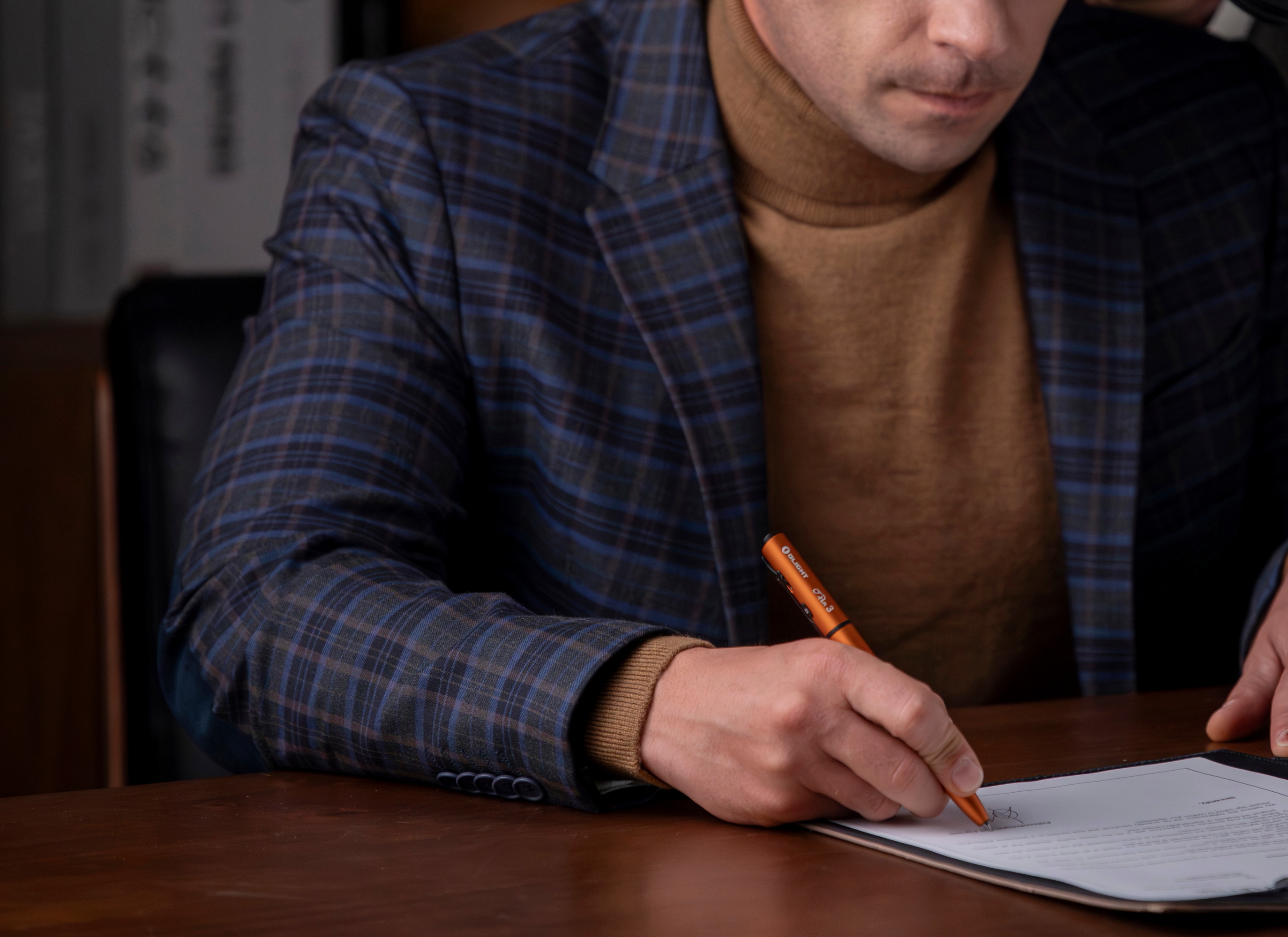 세련된 체크무늬 정장을 입은 남성이 오라이트(Olight) O-Pen 3 주황색 볼펜을 사용하여 중요한 서류에 서명하는 모습