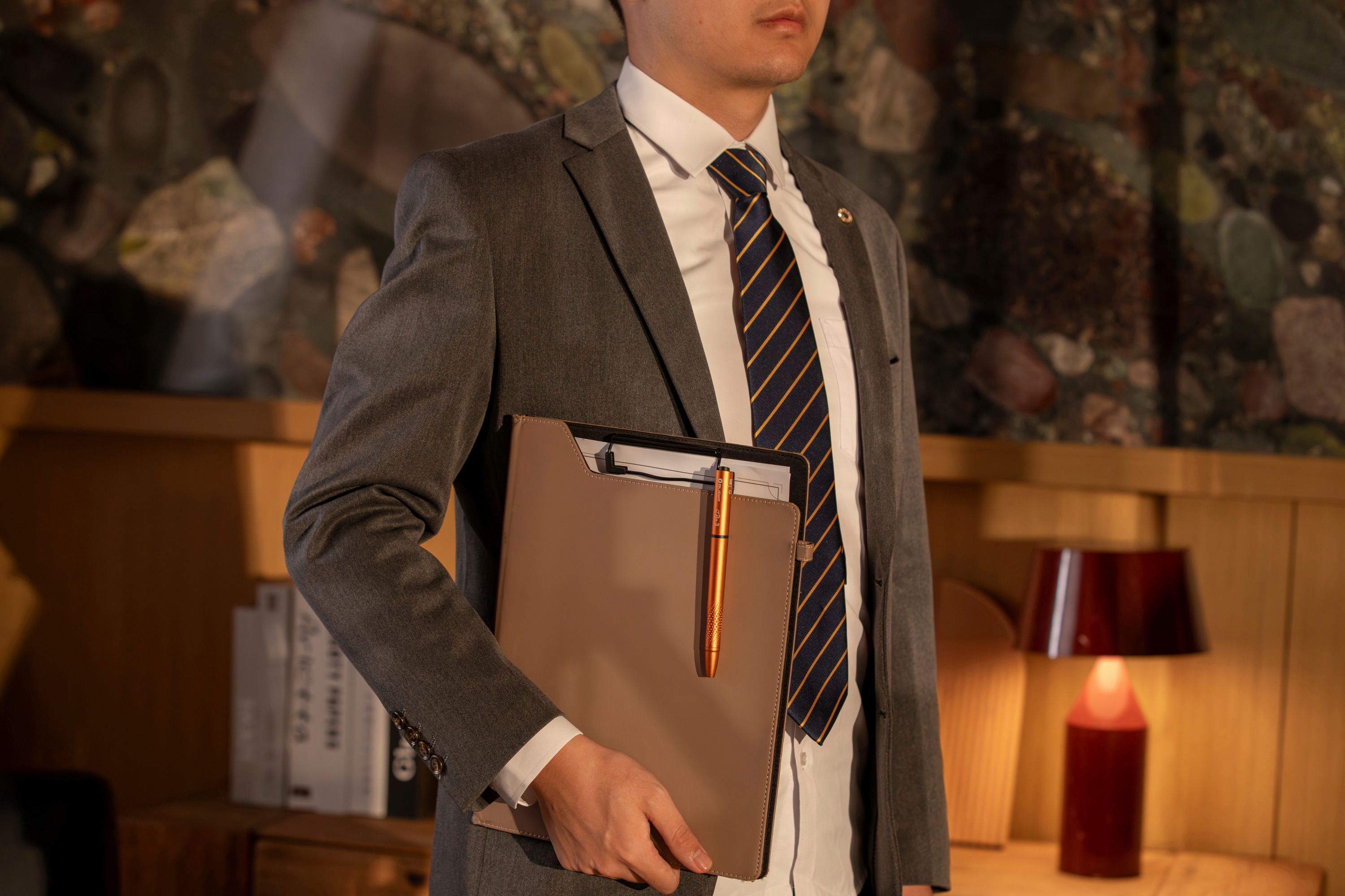 Hombre de negocios con traje gris sosteniendo una carpeta de cuero y un bol&iacute;grafo t&aacute;ctico naranja en un ambiente de oficina profesional. Accesorios elegantes para ejecutivos y moda masculina.
