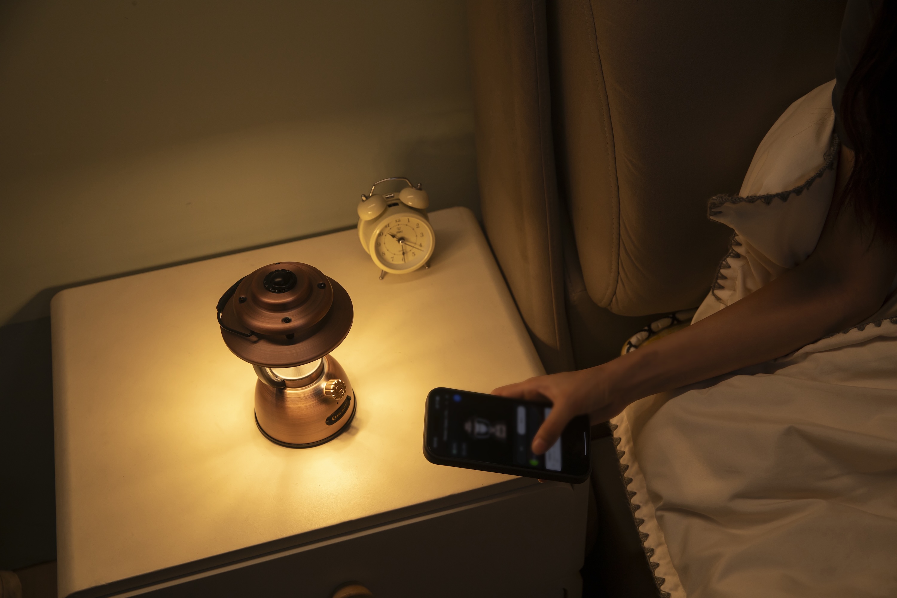 침실 협탁 위 따뜻한 빛을 내는 빈티지 램프와 스마트폰을 조작하는 손.