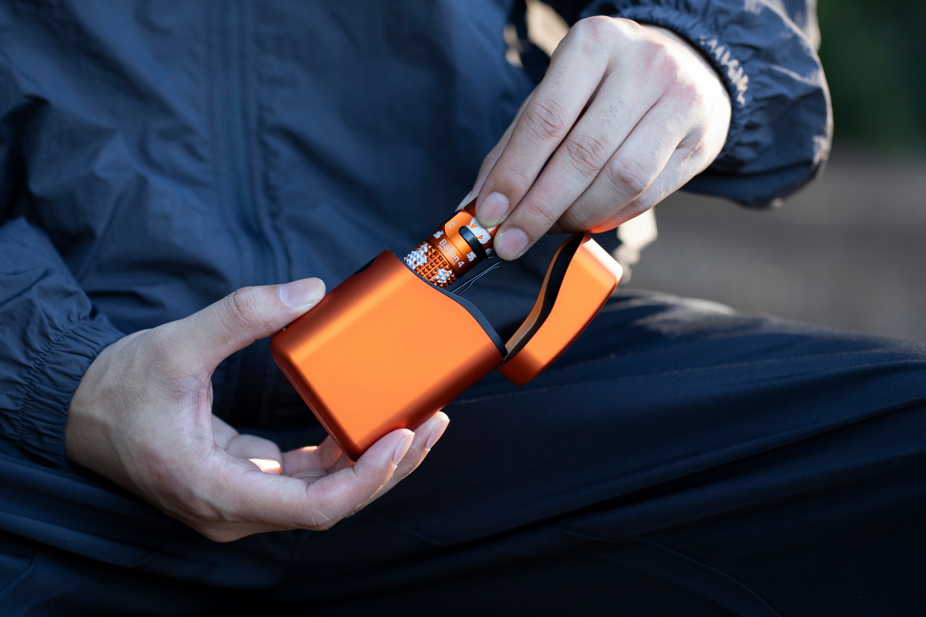 Manos insertando linterna Olight Baton 4 naranja en estuche de carga portátil.