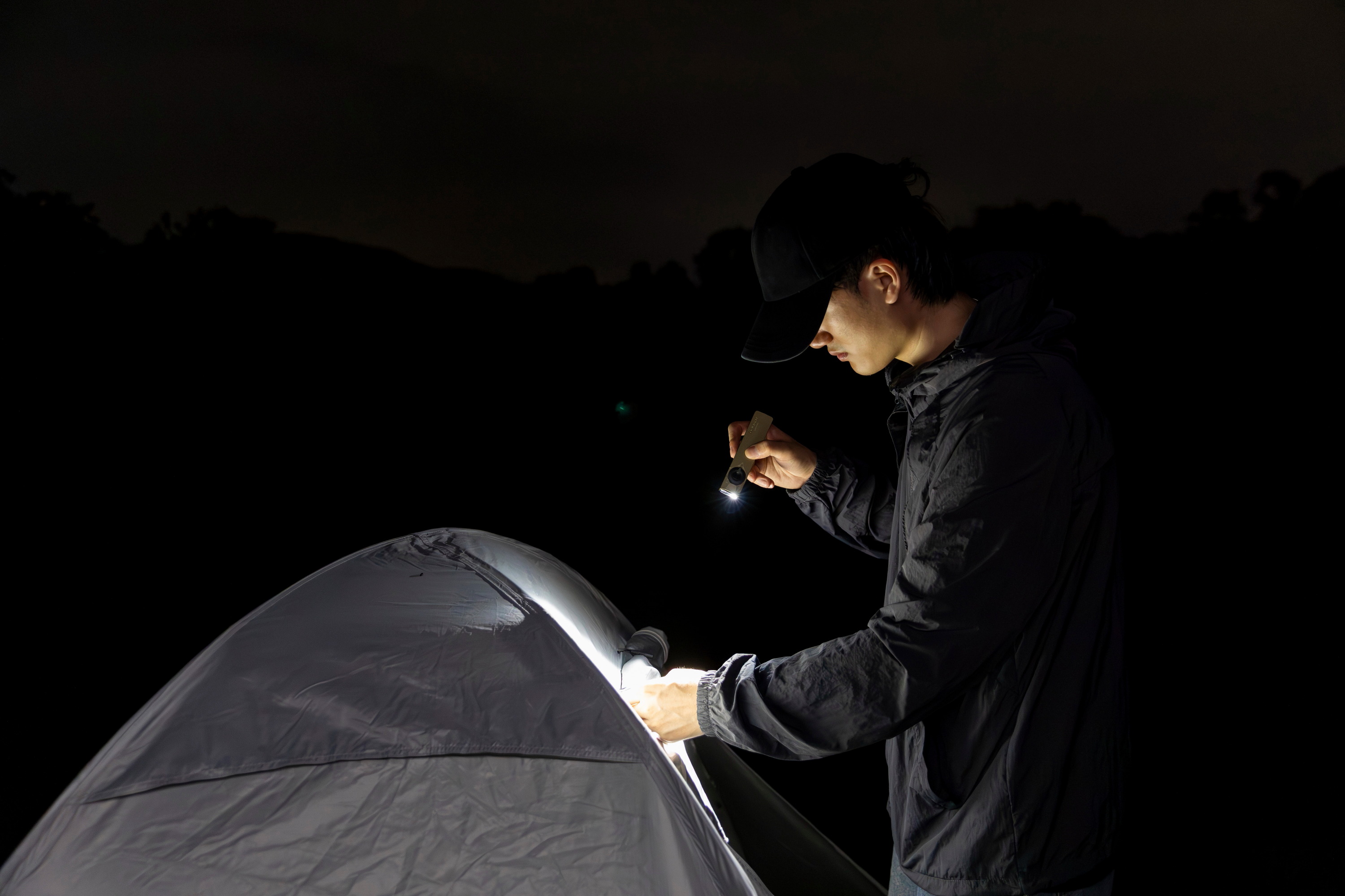 Ein Mann nutzt eine flache LED-Taschenlampe beim Camping, um nachts sein Zelt zu beleuchten. Kompakte Outdoor-Beleuchtung für Wanderungen und Campingausflüge bei Dunkelheit.