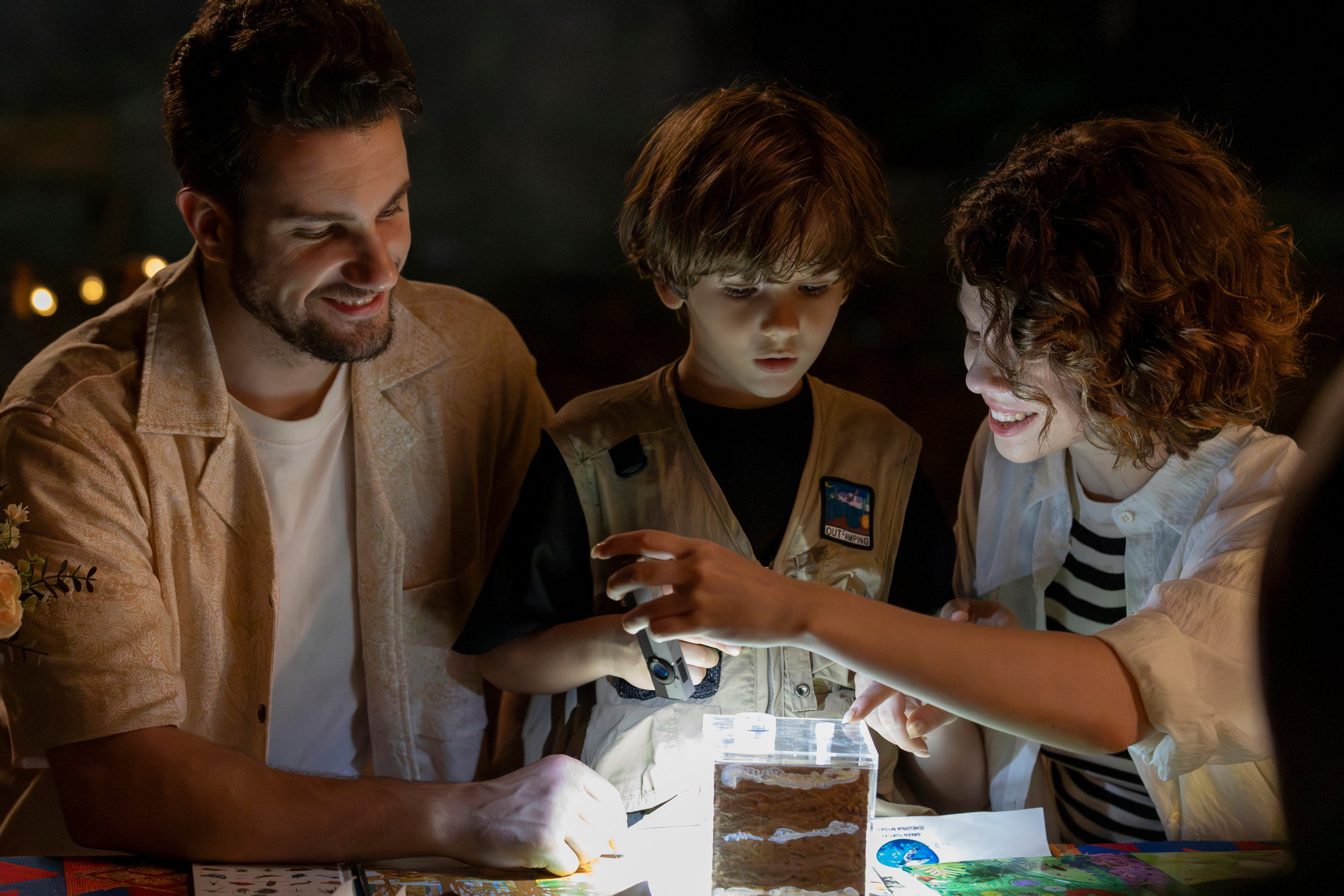 Happy family with a young boy using a magnifying glass and flashlight to examine insects in a container during a night camping science activity.