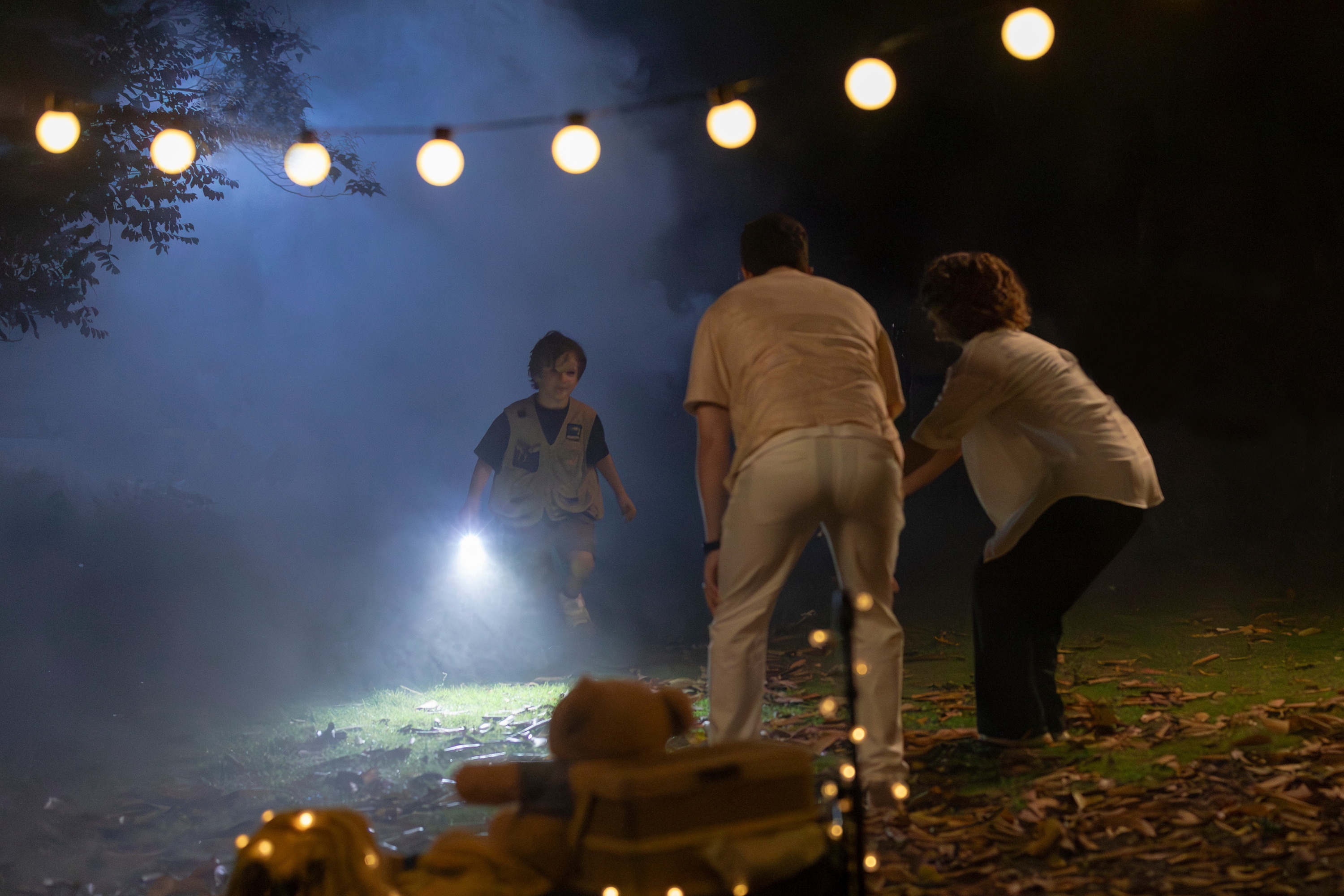 Enfant scout avec lampe de poche explorant un jardin brumeux la nuit, rejoignant deux adultes sous des guirlandes lumineuses. Sc&egrave;ne d'aventure nocturne et de camping en famille dans une ambiance myst&eacute;rieuse et ludique.