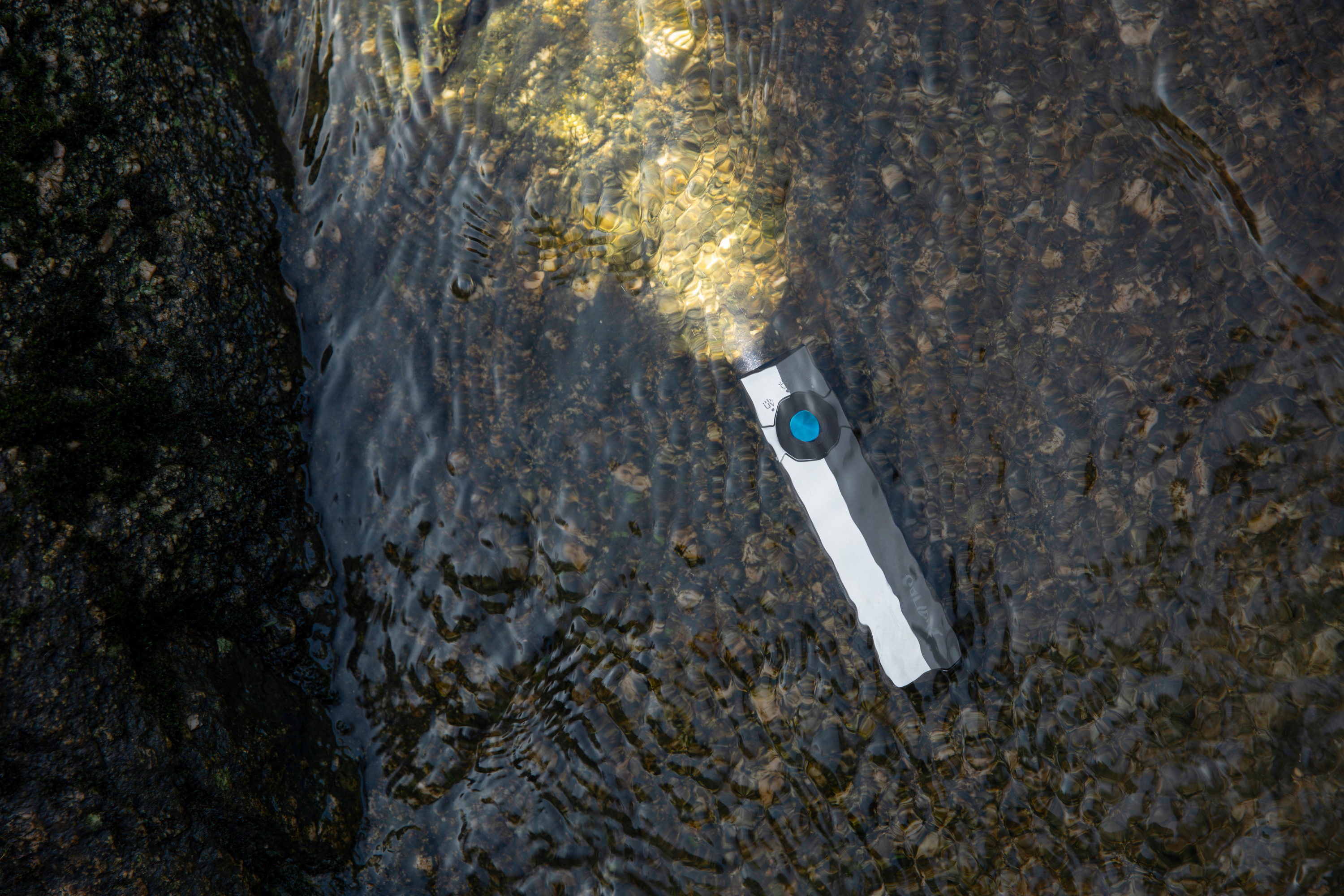 Olight ArkPro flat flashlight with a blue button submerged in water, showing a bright beam of light reflecting off river stones.