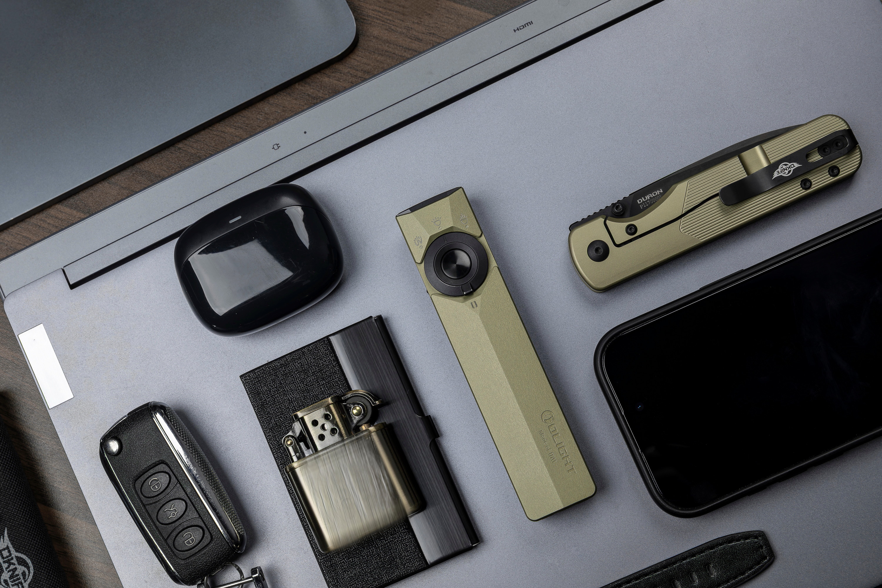 Everyday carry EDC gear flat lay featuring olive green Olight Arkfeld flashlight, Oknife pocket knife, vintage lighter, car key fob, wireless earbuds, and smartphone on a grey laptop surface.