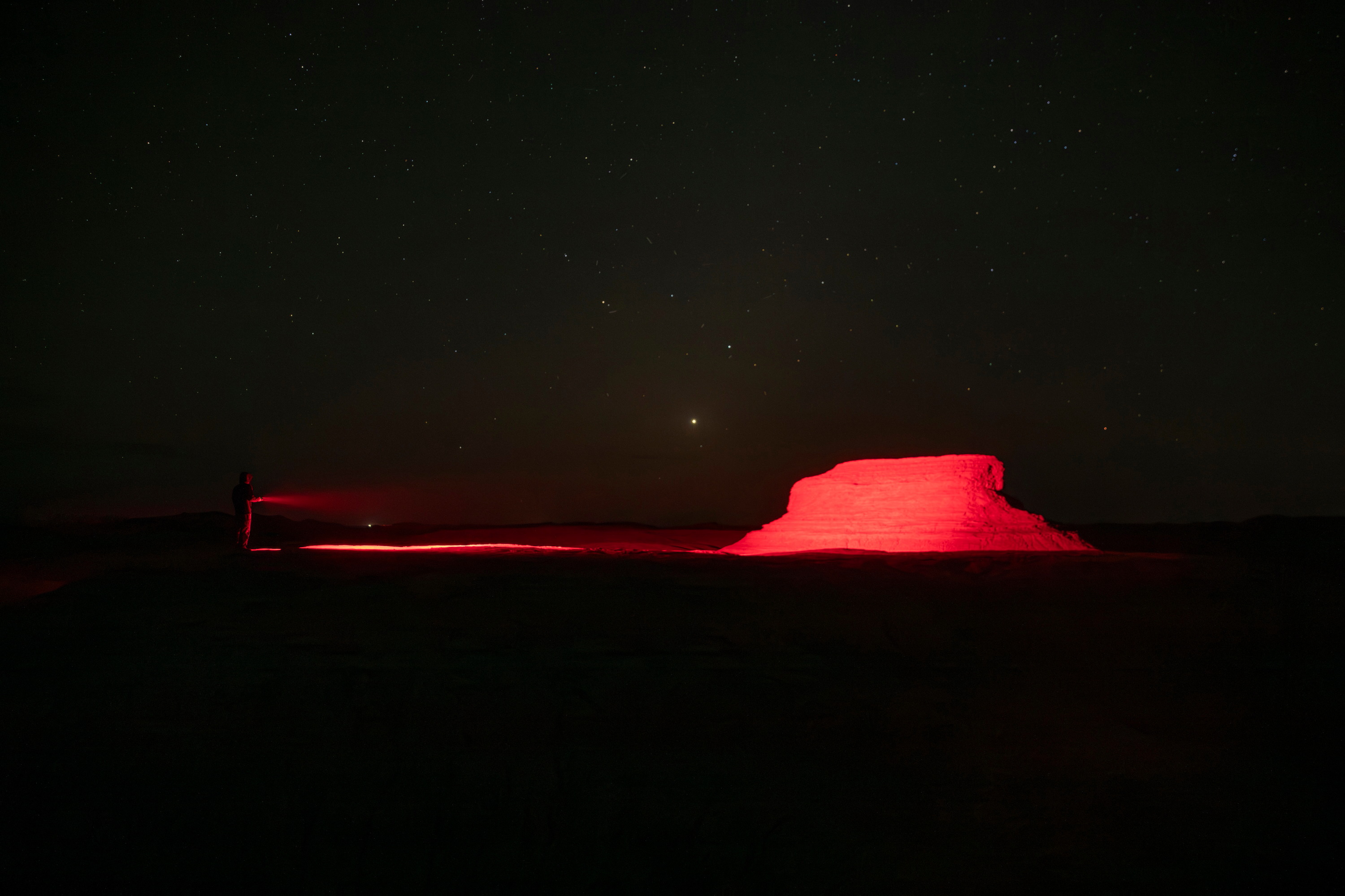 Persona iluminando formación rocosa con luz roja bajo un cielo nocturno estrellado en el desierto.