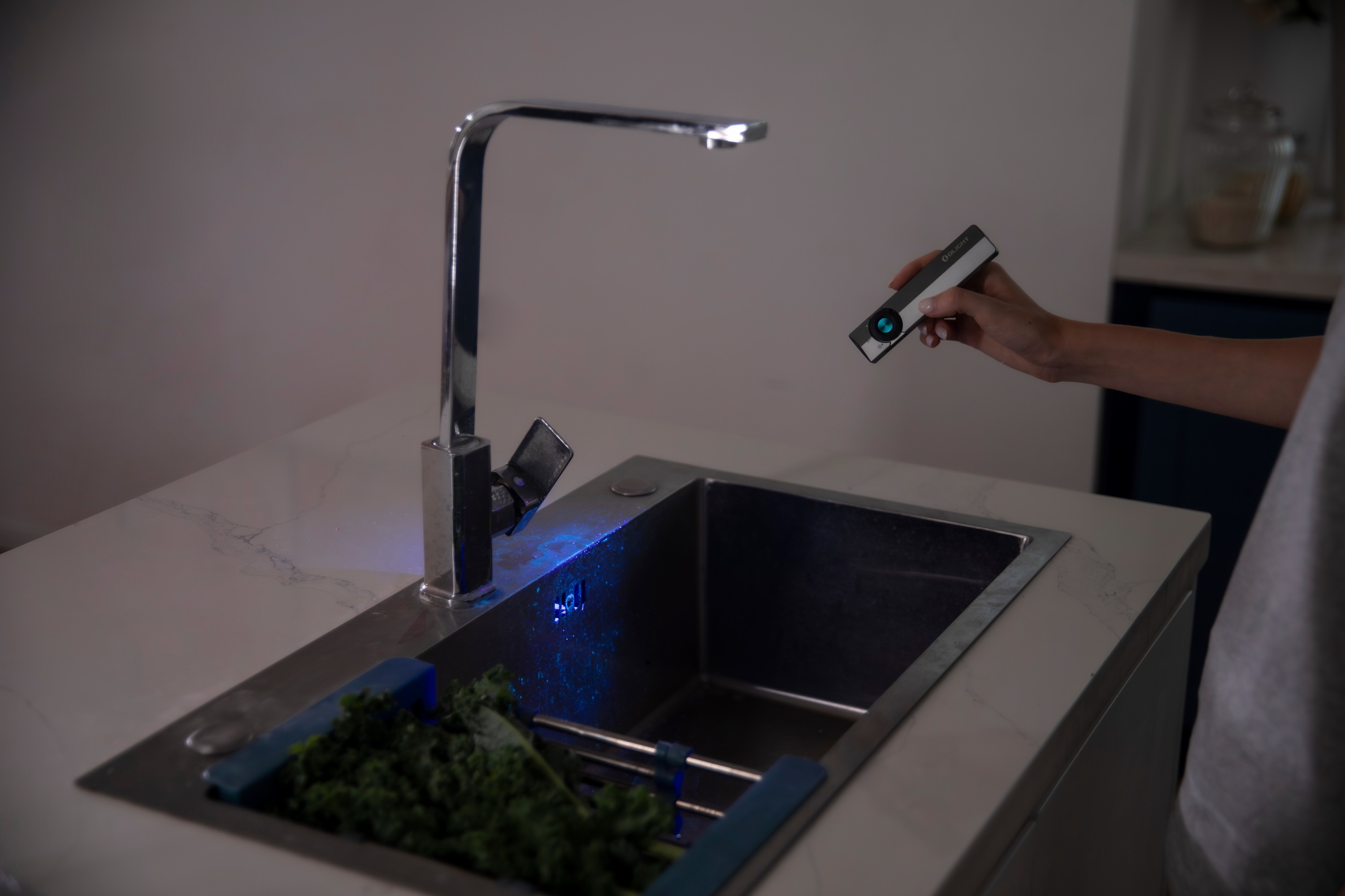 Hand using a UV light to inspect a kitchen sink with green vegetables, revealing cleanliness or residue for hygiene purposes.